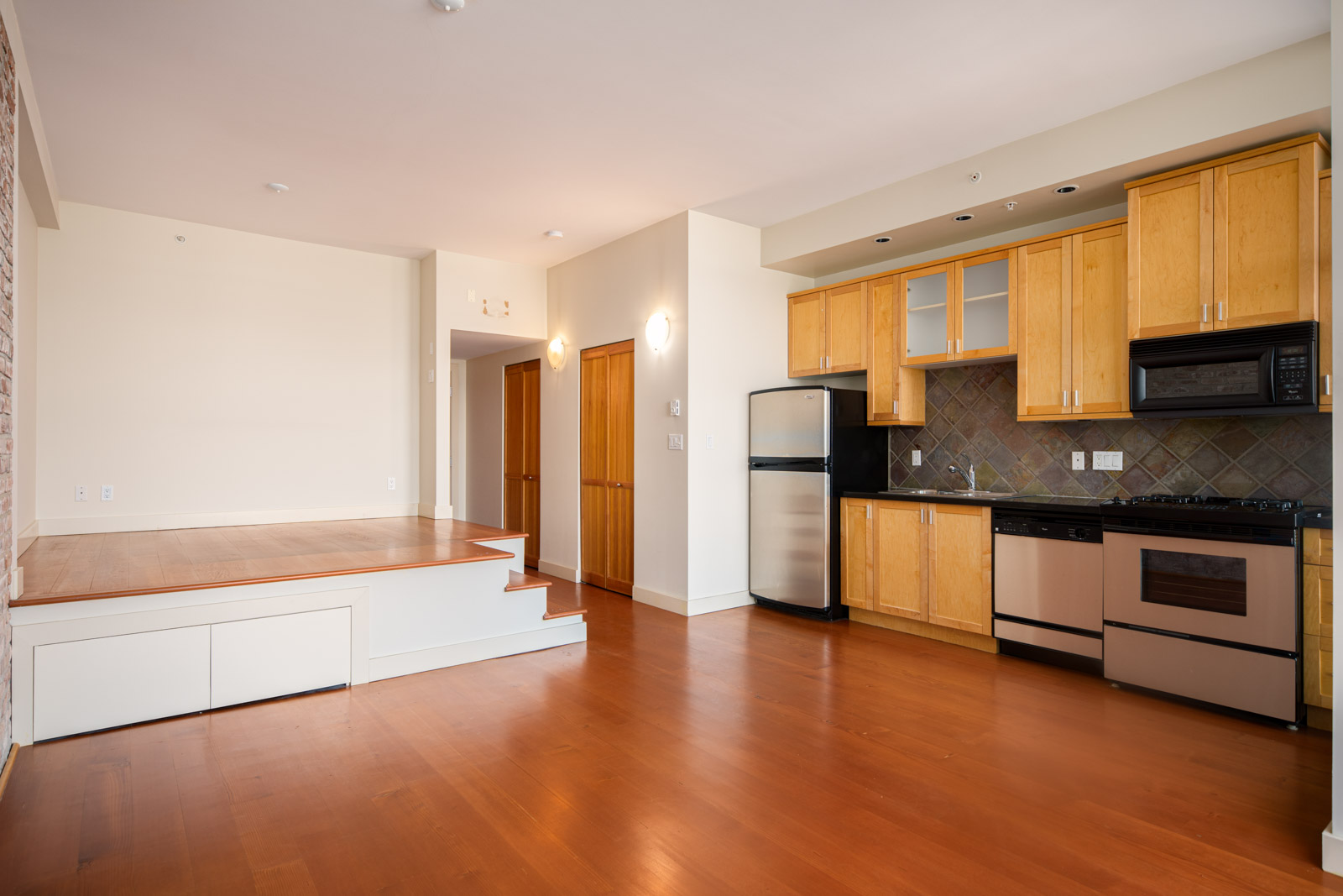 Modern apartment with wood flooring, a small raised platform, and an open kitchen featuring stainless steel appliances and light wood cabinets.