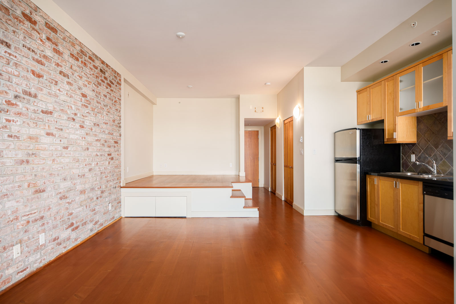 Open-plan apartment with hardwood floors, exposed brick accent wall, elevated platform area, and a kitchen featuring wood cabinets and stainless steel appliances.