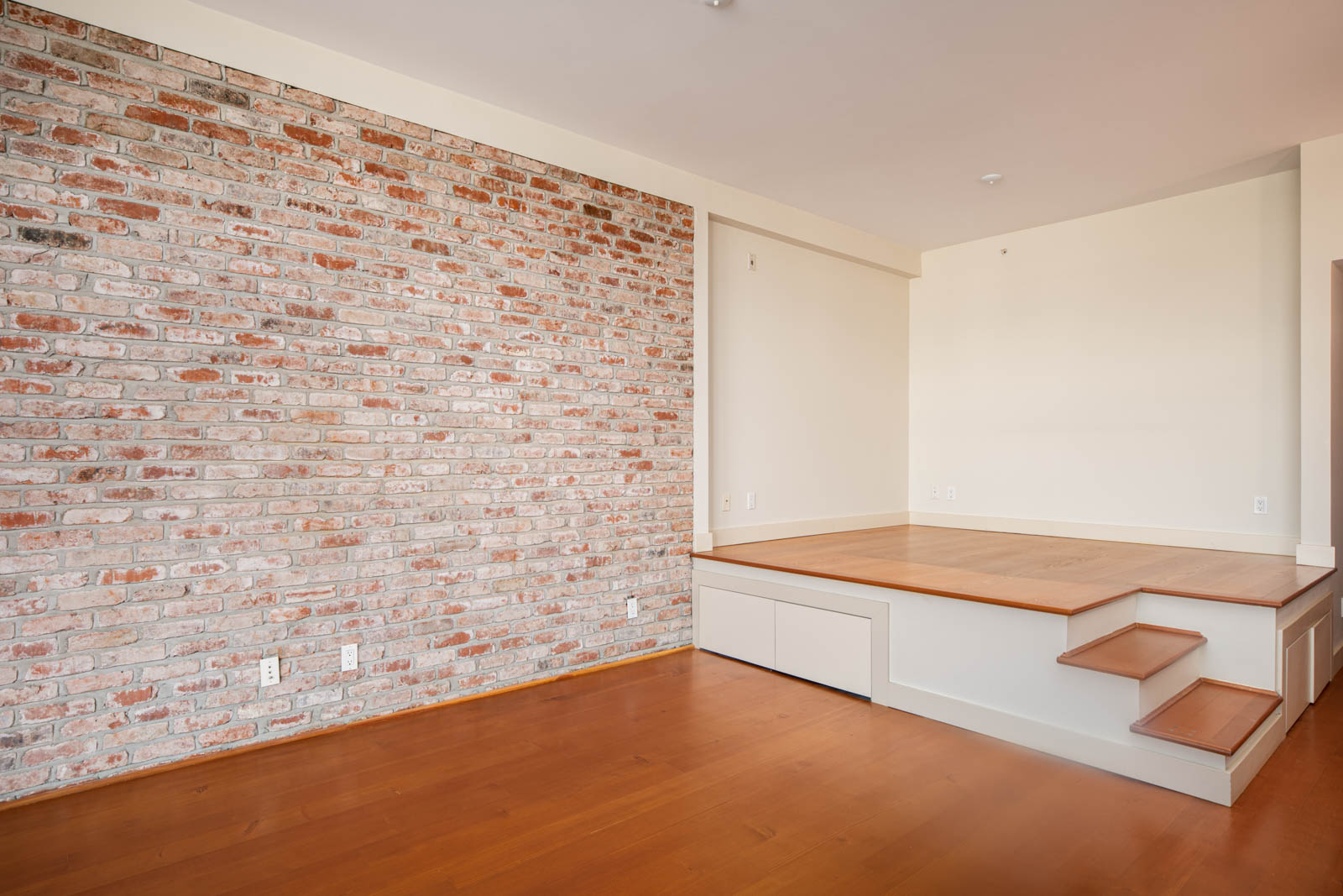 Room with a brick accent wall, raised wooden platform with built-in drawers, small steps, and hardwood floors. Walls are painted light beige.