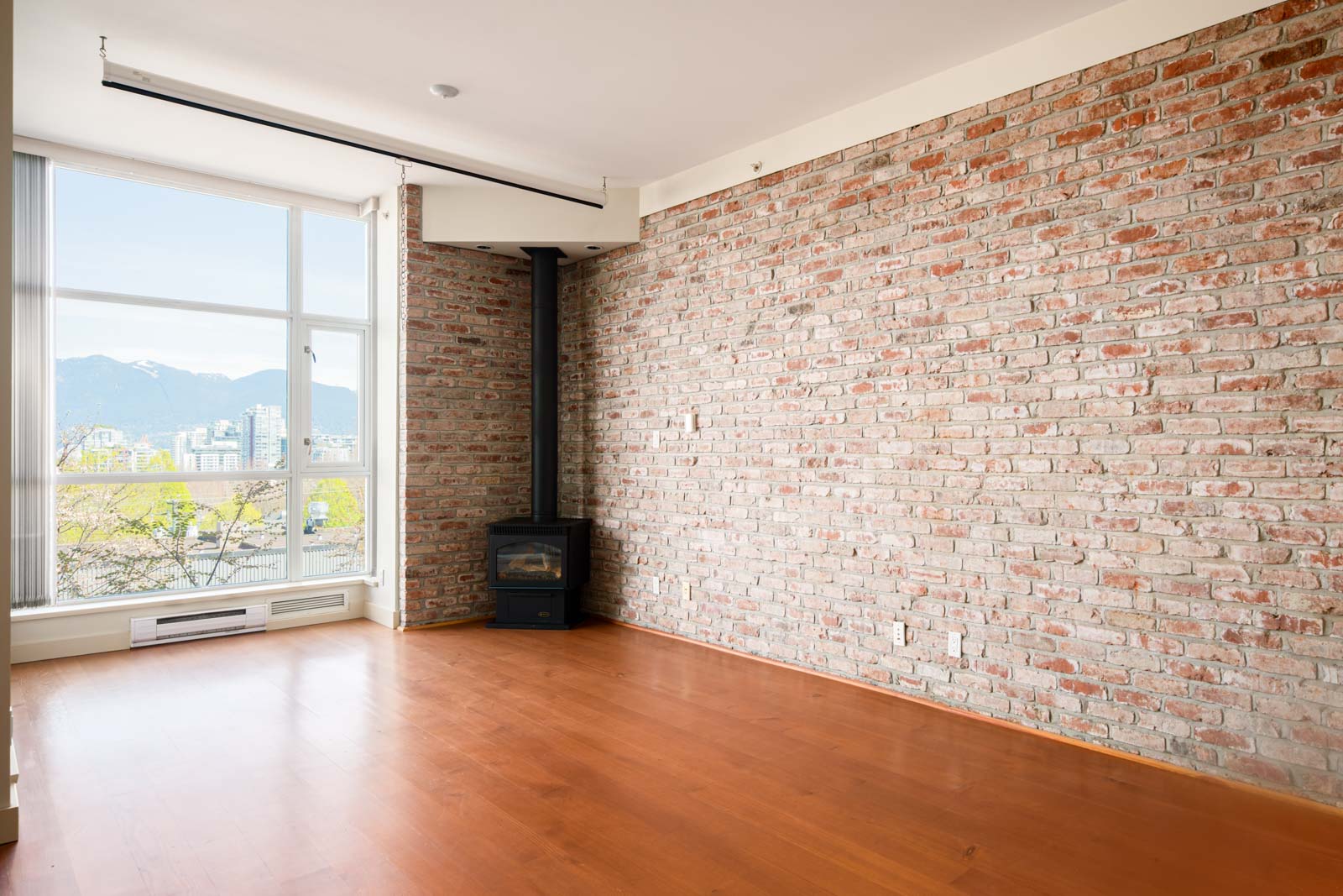 Empty room with exposed brick wall, hardwood floor, a wood stove in the corner, and large windows offering a view of buildings and mountains.