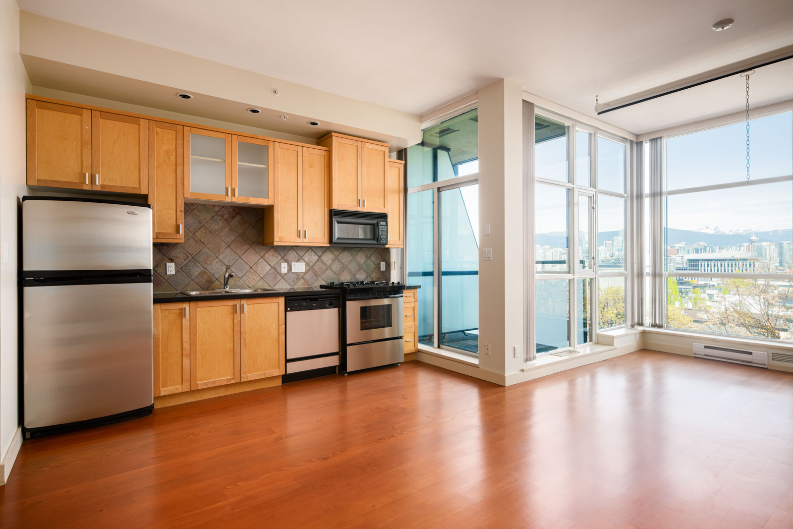 Modern apartment kitchen with light wood cabinets, stainless steel appliances, and large floor-to-ceiling windows overlooking a cityscape.
