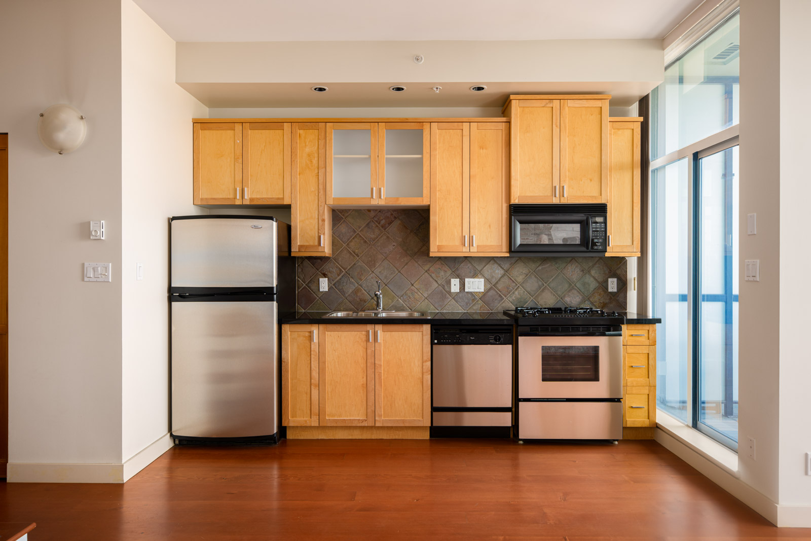 Modern kitchen with wooden cabinets, stainless steel fridge, dishwasher, oven, gas stove, and microwave, set against a tiled backsplash and hardwood floor.