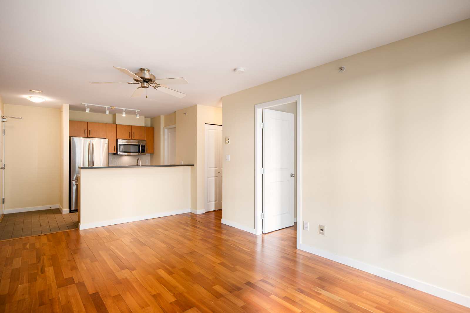 Empty modern apartment interior with hardwood floors, beige walls, a kitchen area with stainless steel appliances, and a ceiling fan.