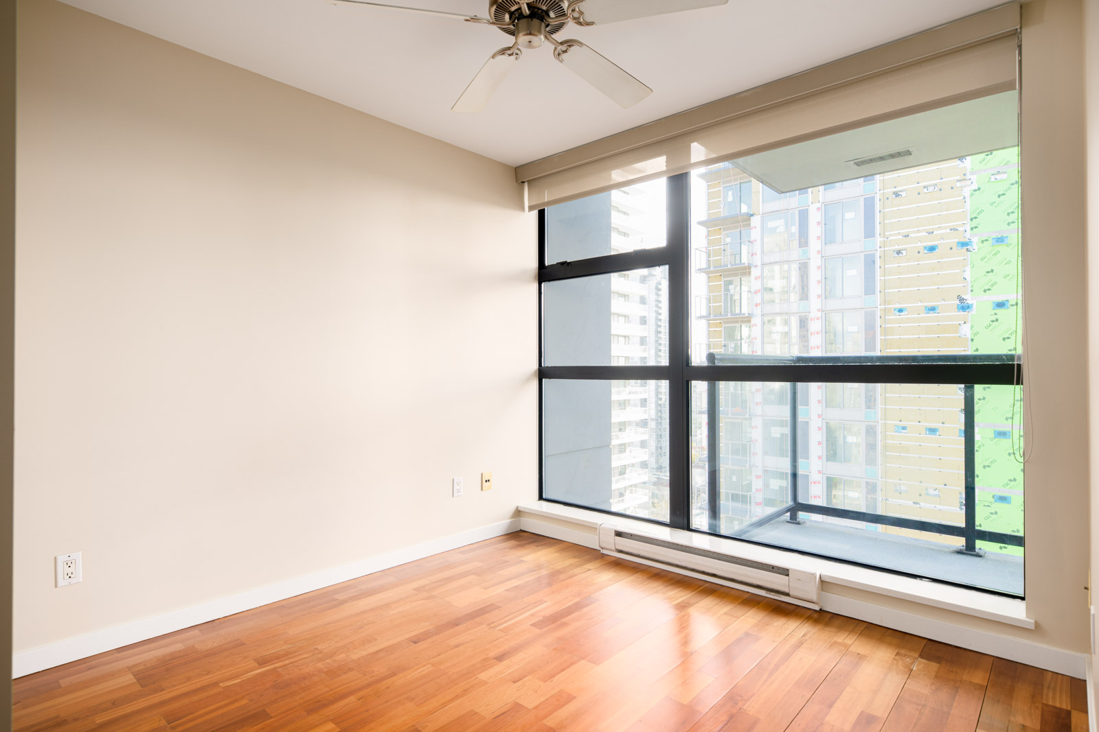 Empty room with beige walls, hardwood floors, a ceiling fan, and large floor-to-ceiling windows overlooking a building under construction.