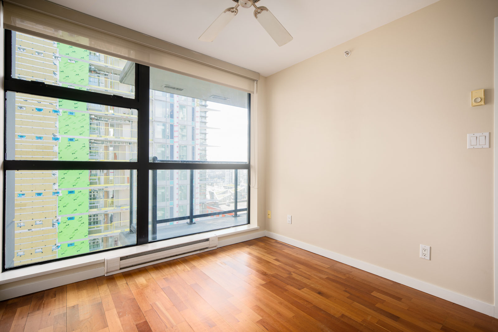 Empty room with light beige walls, hardwood floor, ceiling fan, large window, and view of nearby construction on a tall building.