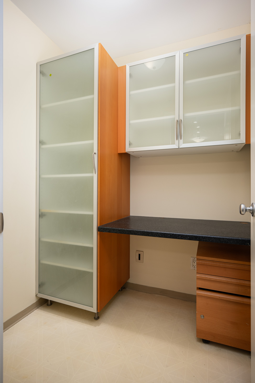 Small room with built-in wooden cabinets, frosted glass doors, a black countertop, and a rolling drawer unit, all set against cream-colored walls and flooring.