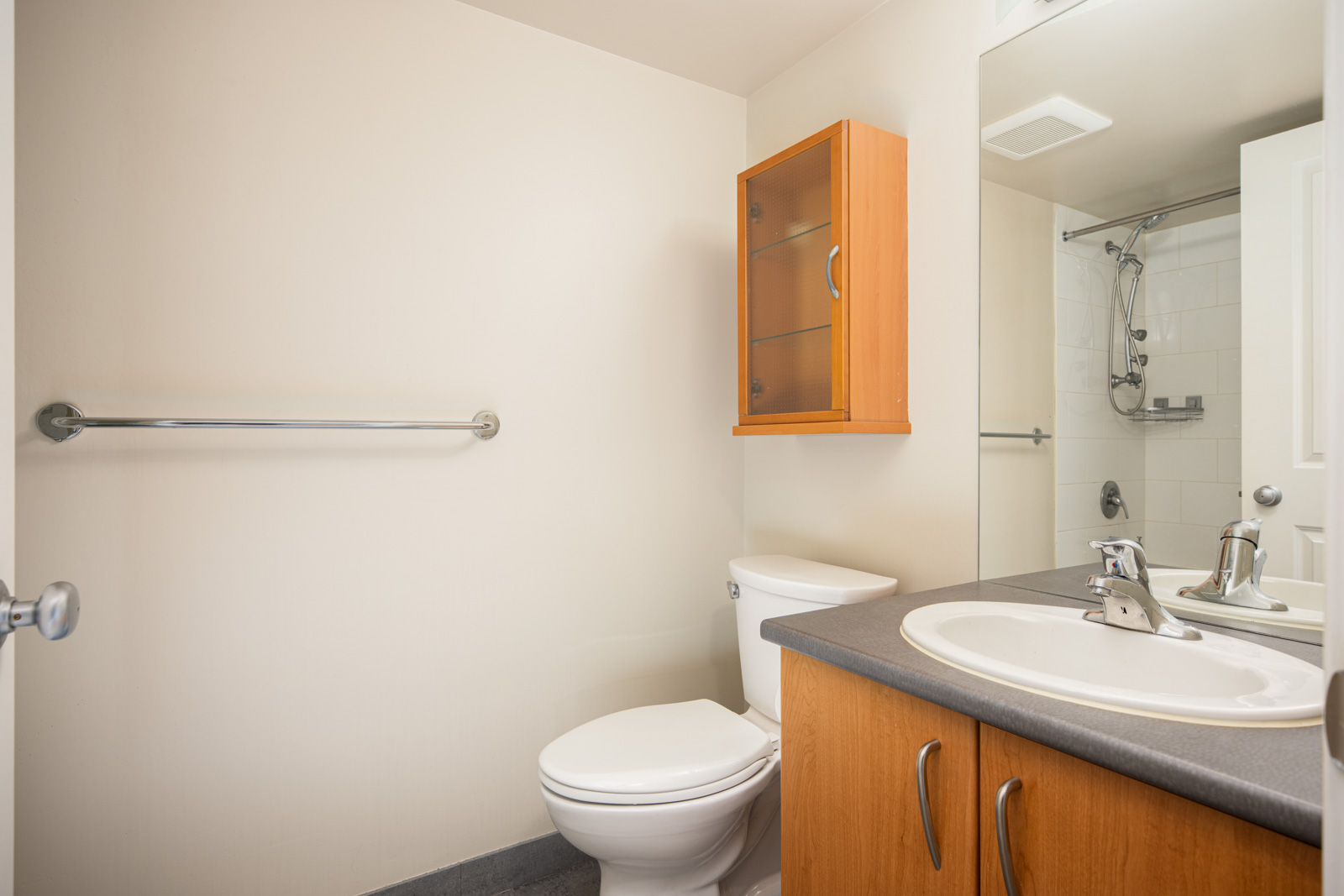 A bathroom with a sink, countertop, toilet, wall-mounted cabinet, towel bar, mirror, and a shower with a glass door.