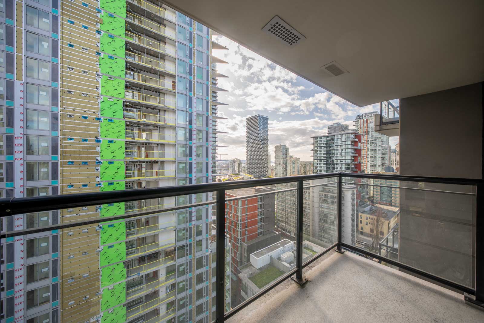 View from a high-rise balcony overlooking nearby construction and modern tall buildings in an urban cityscape under a partly cloudy sky.