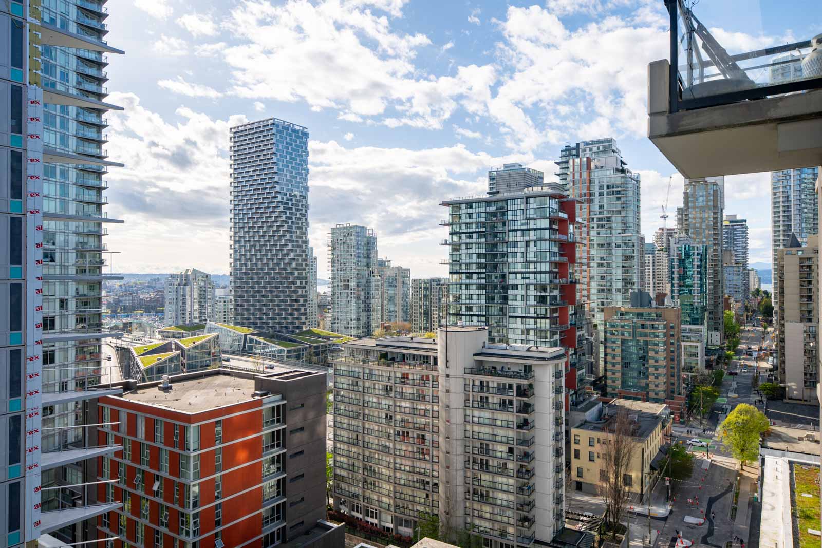 View of a modern city skyline with multiple high-rise apartment buildings, glass towers, and a few trees, under a partly cloudy sky.