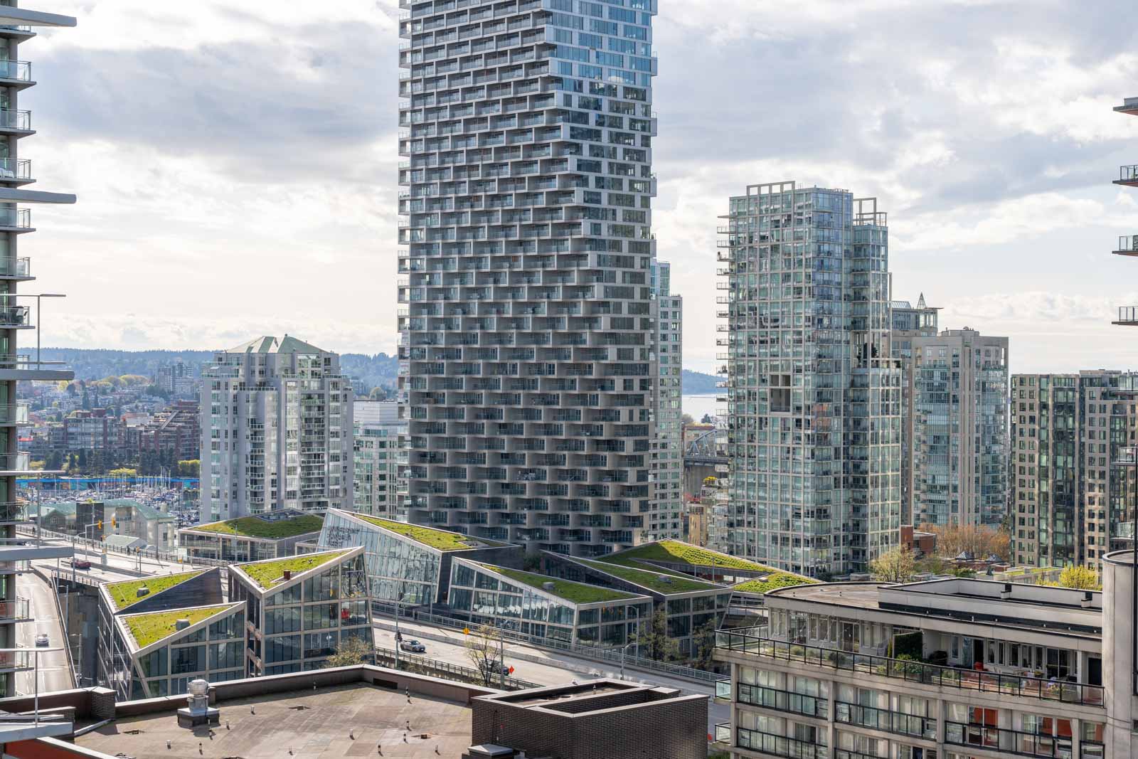 Cityscape featuring modern high-rise buildings with geometric facades and rooftop greenery, under a cloudy sky. A few roads and partial waterfront are visible in the background.