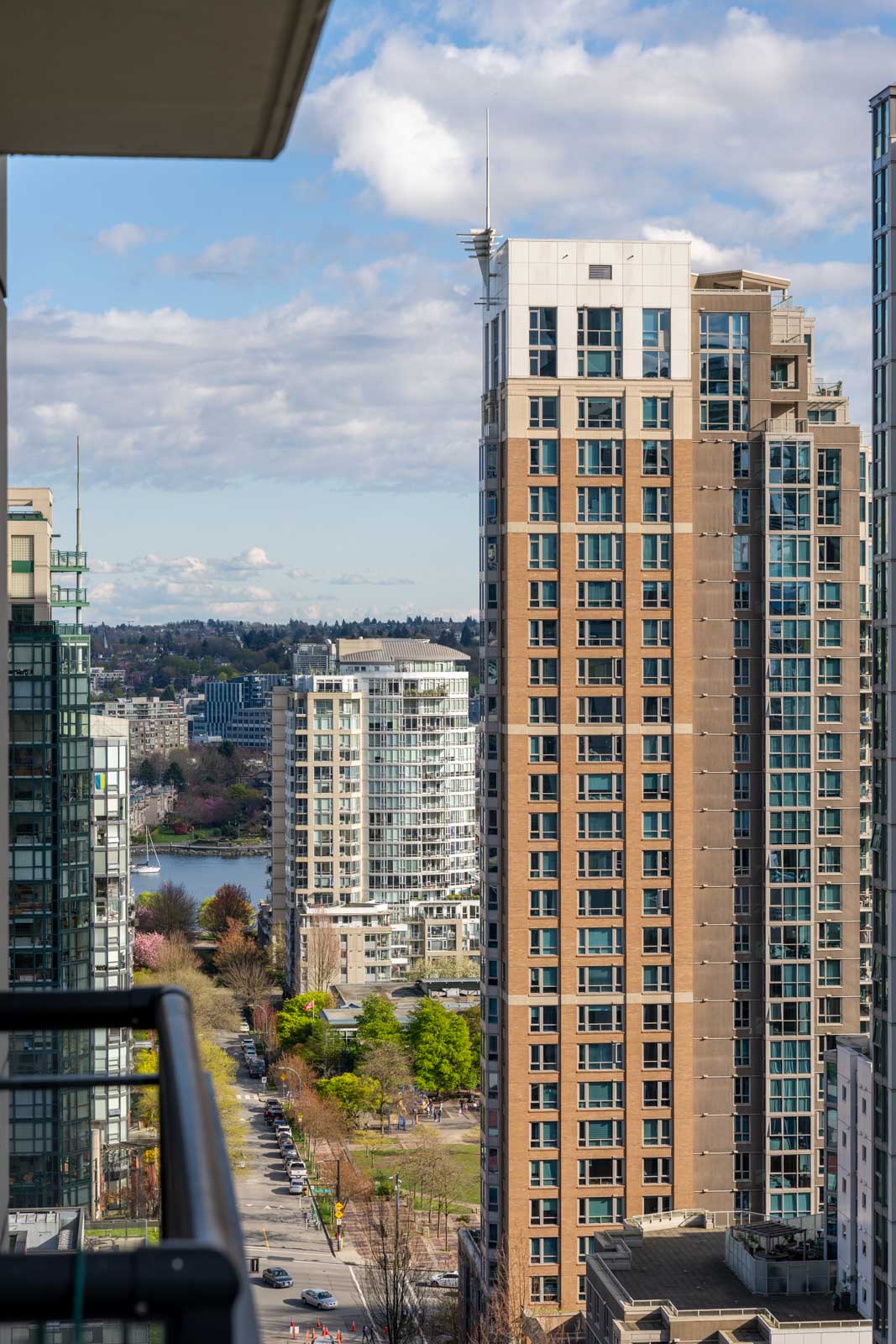 View from a high-rise balcony showing tall residential buildings, a tree-lined street below, and a river with more buildings in the distance under a partly cloudy sky.