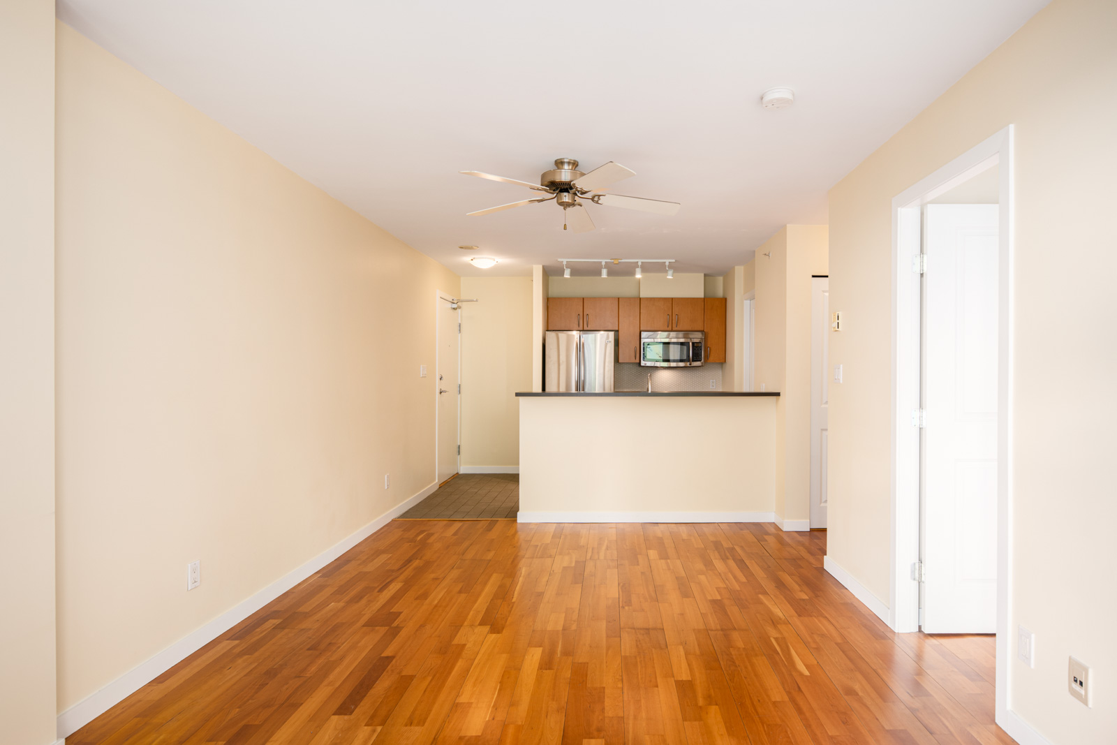 Unfurnished apartment interior with hardwood floors, beige walls, ceiling fan, open kitchen with stainless steel appliances, and white doors.