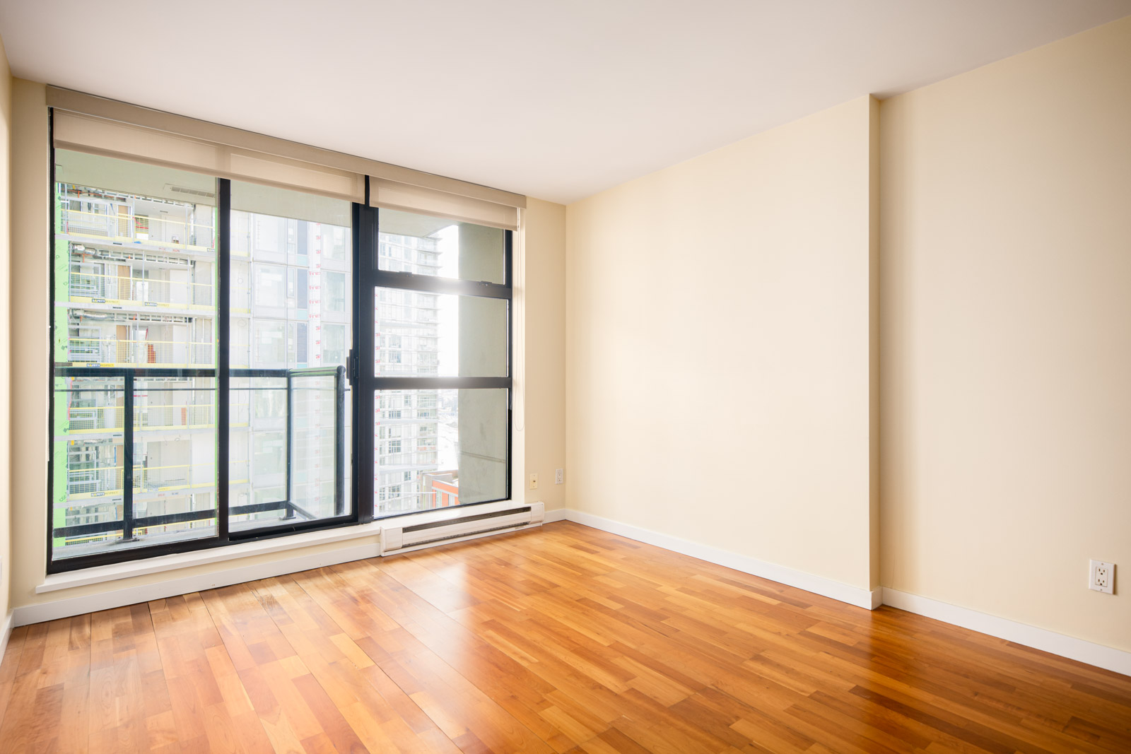Empty room with light-colored walls, wood flooring, and a large floor-to-ceiling window showing city buildings outside.