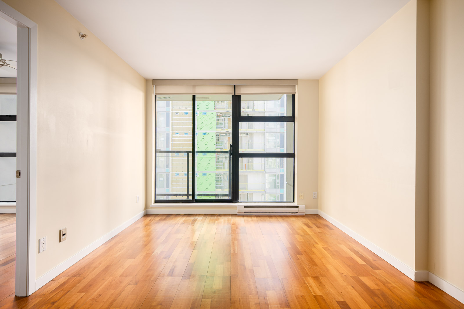 Empty room with light-colored walls, large floor-to-ceiling windows, and wooden floors; adjacent open doorway visible on the left.