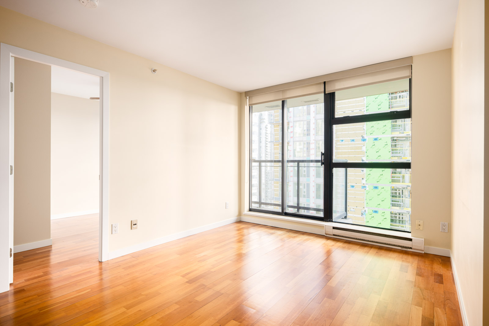 Unfurnished room with light-colored walls, large window with city view, wooden floor, and an open doorway leading to another room.