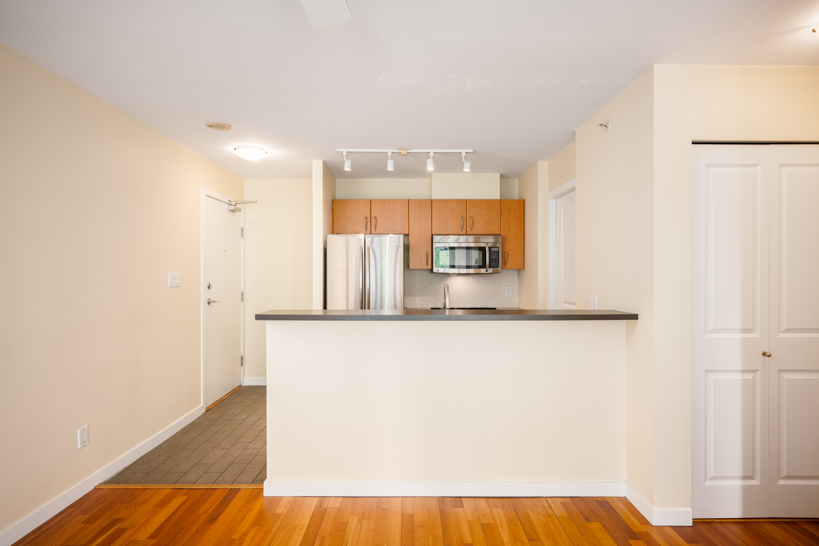 A modern kitchen with stainless steel appliances, wood cabinets, a breakfast bar, and track lighting, adjacent to an entryway with tile flooring.