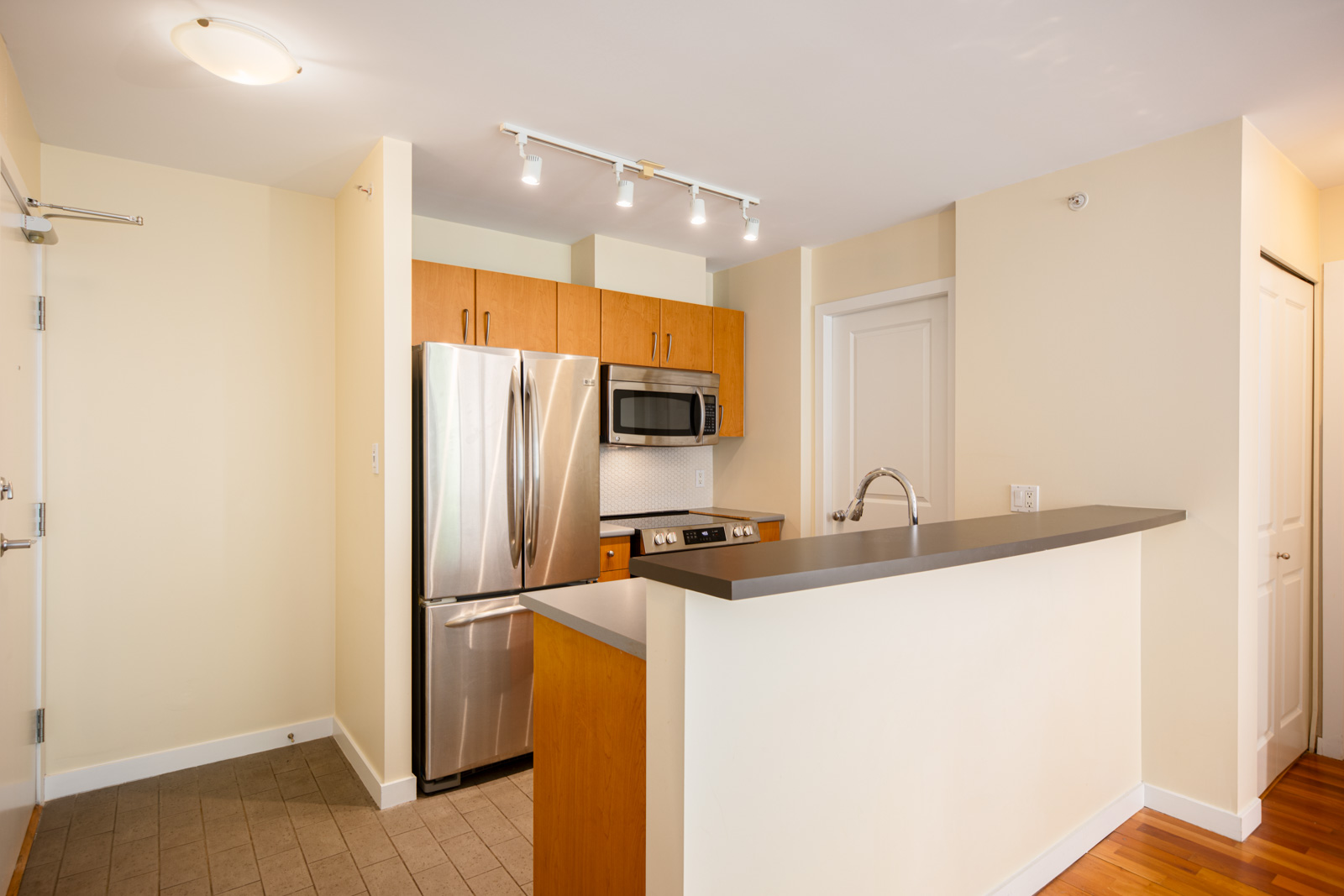 Modern kitchen with stainless steel appliances, wood cabinets, a microwave, a refrigerator, a sink, and a breakfast bar with a dark countertop. Cream-colored walls and wood flooring.