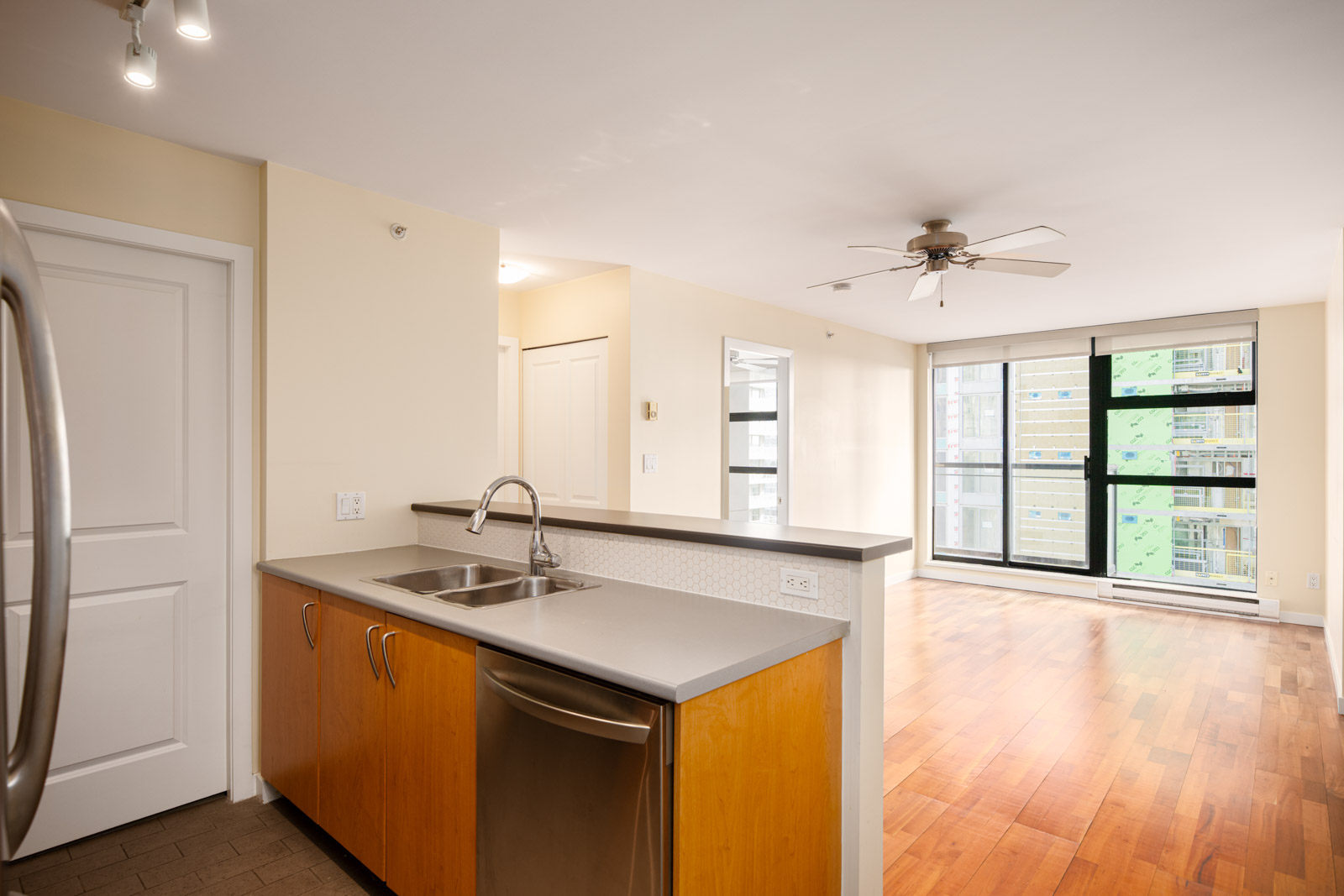 A modern kitchen with a double sink and dishwasher opens to a bright living area with wood flooring, large windows, and a ceiling fan.