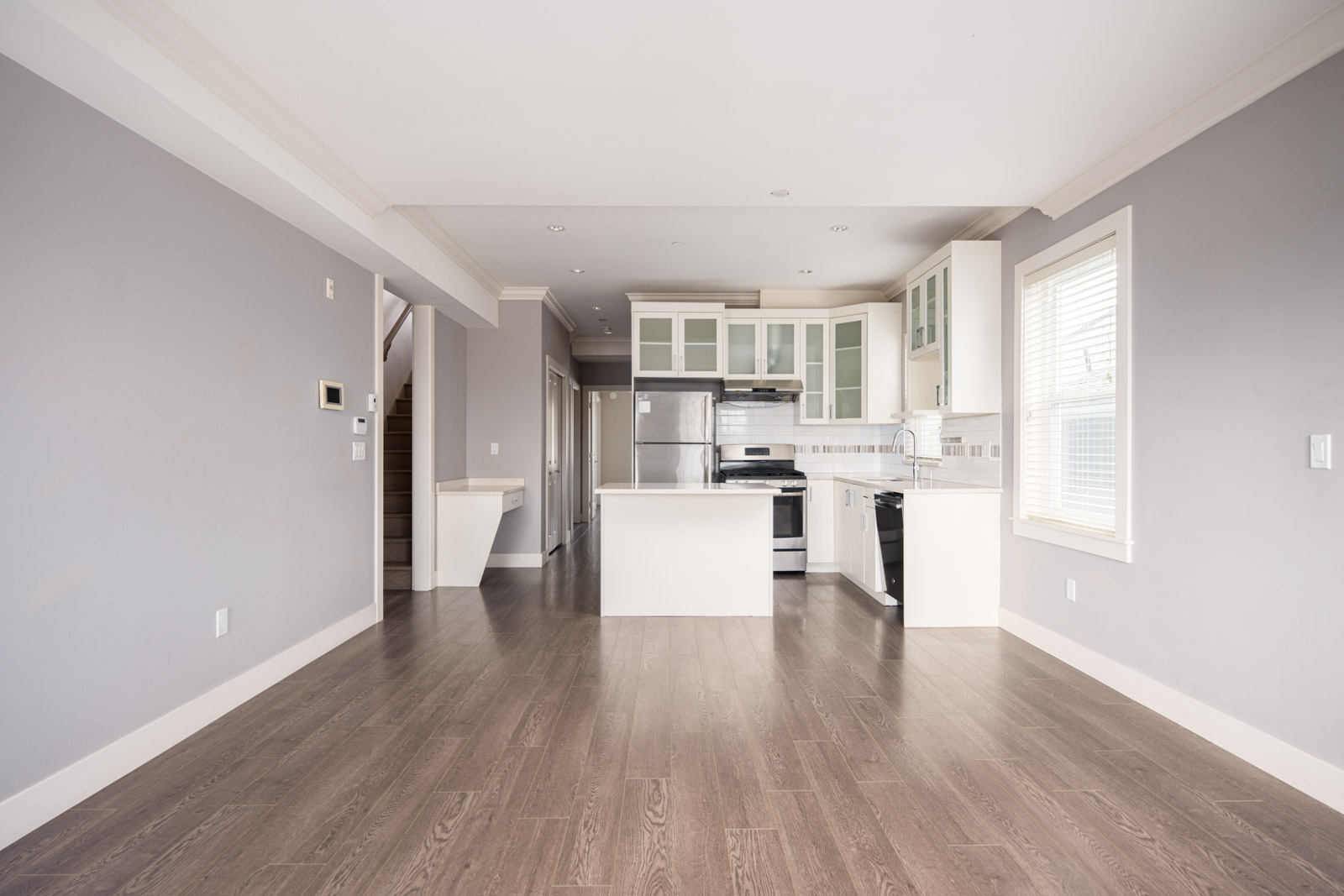 A modern, empty kitchen and living area with gray walls, wood flooring, stainless steel appliances, and white cabinetry. Natural light comes in through a window on the right.