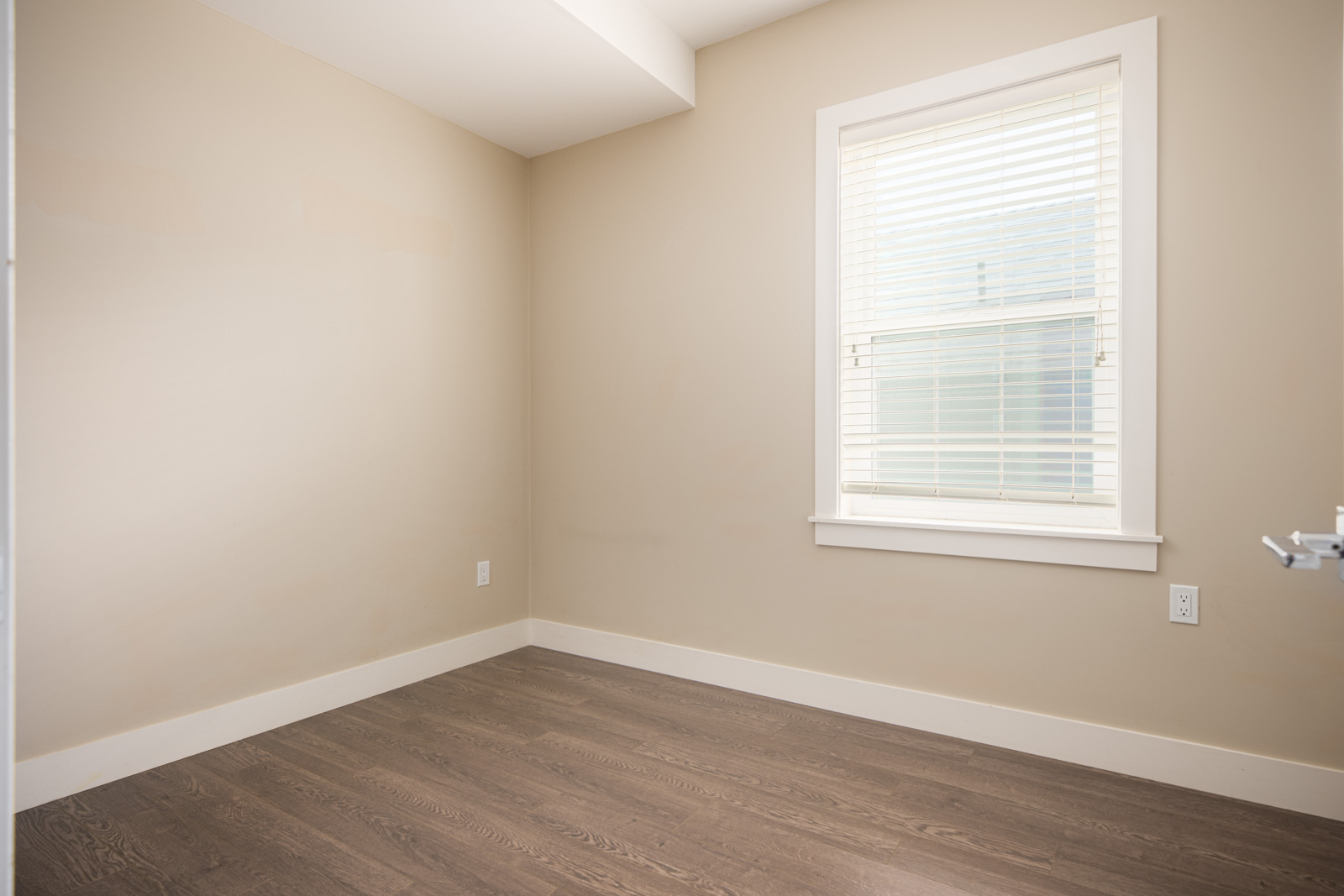 Empty room with beige walls, a single window with white blinds, white trim, and dark wood flooring.