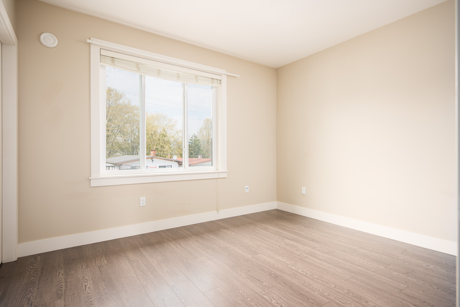 Empty room with beige walls, a large window with white blinds, and light brown wood flooring. Daylight comes in through the window, showing houses and trees outside.