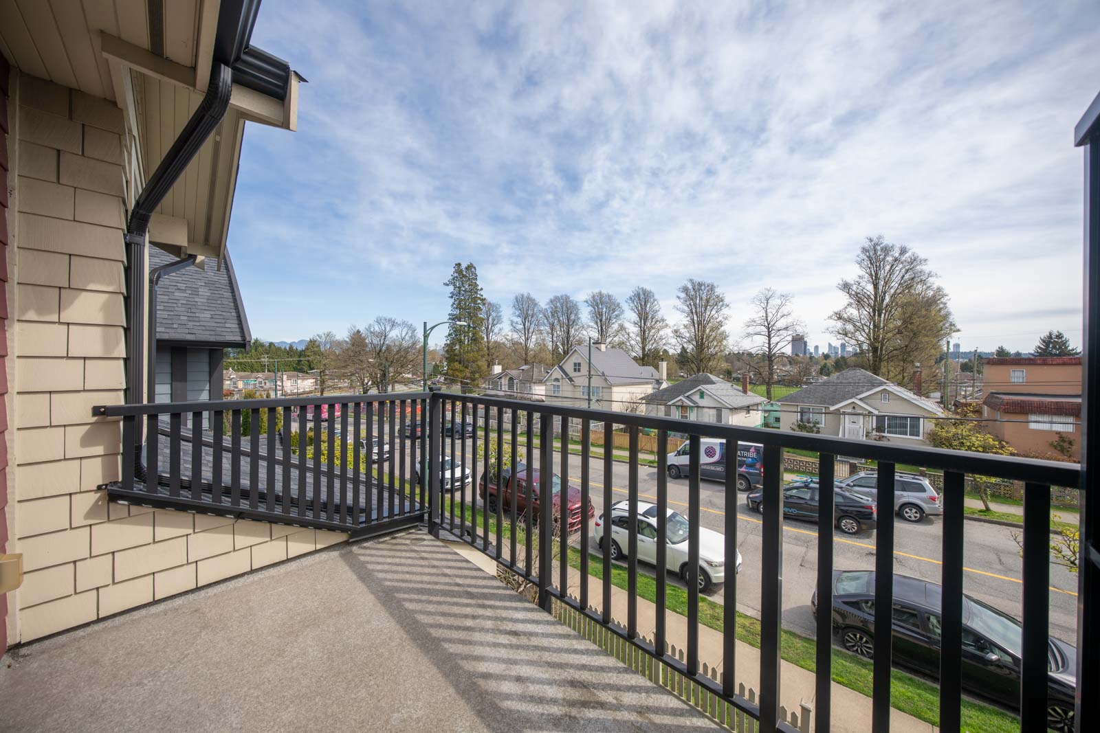 View from a balcony overlooking a residential street with parked cars, houses, trees, and a partly cloudy sky in the background.