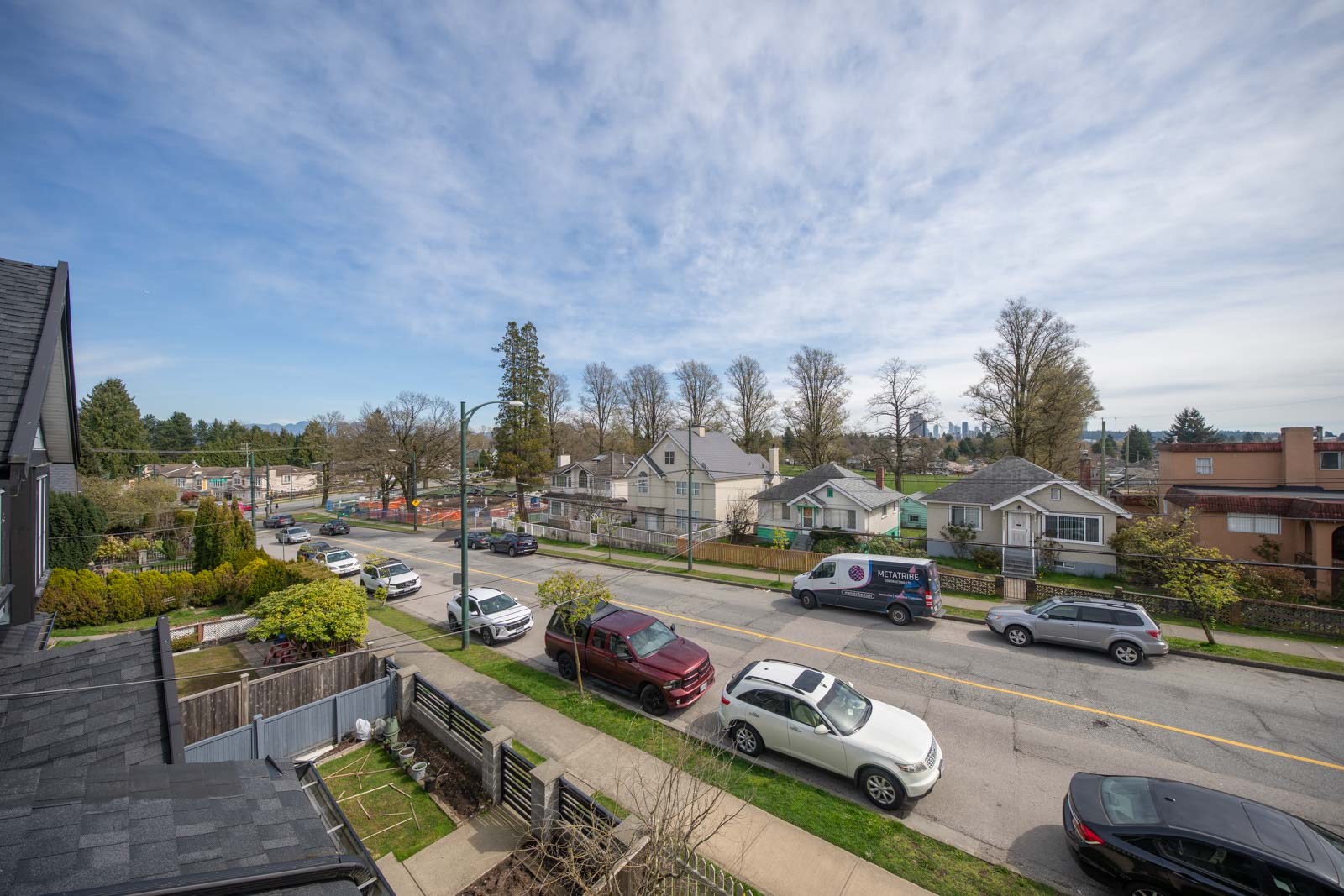 Residential street with parked cars, houses, trees without leaves, and a partly cloudy sky seen from an elevated perspective.