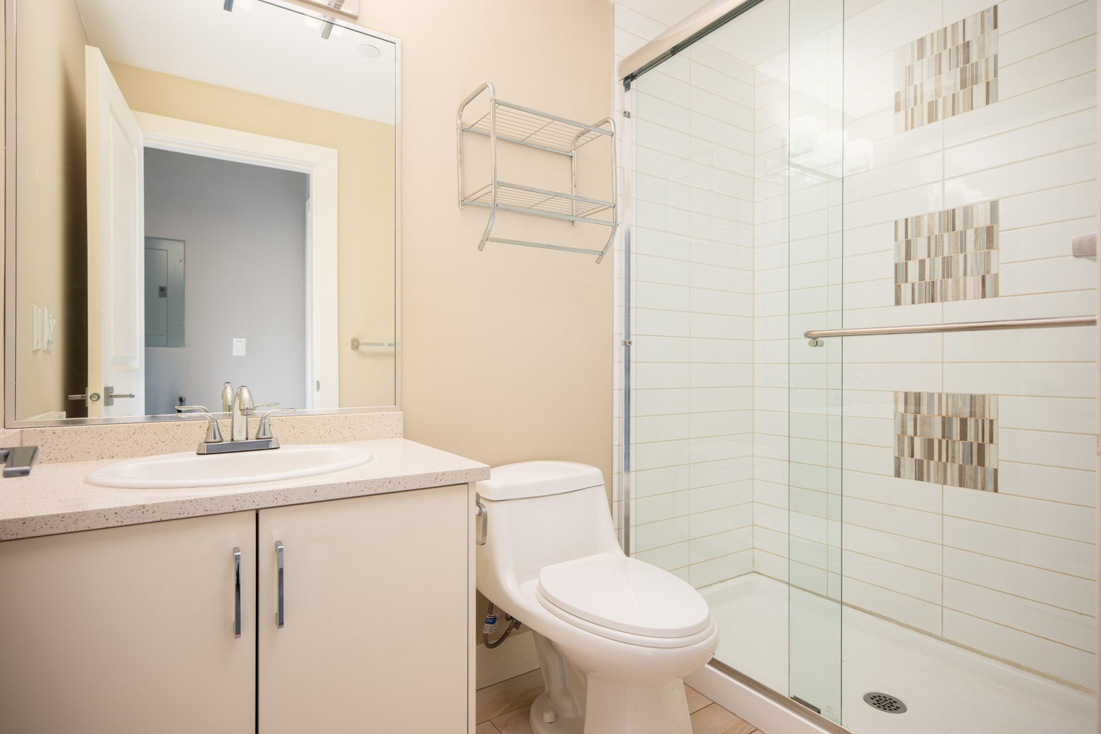 Modern bathroom with a white vanity, integrated sink, toilet, wall-mounted shelf, and a glass-enclosed shower with decorative tile accents.