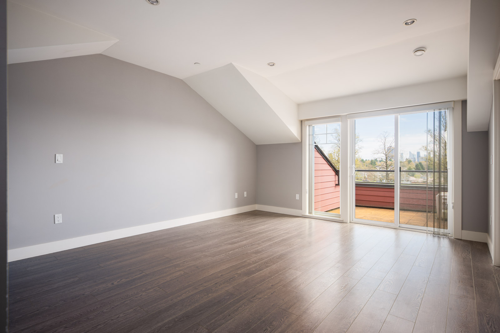 Empty room with light gray walls, dark wood floors, sloped ceiling, and large glass doors leading to a balcony with a view of trees and city buildings in the distance.
