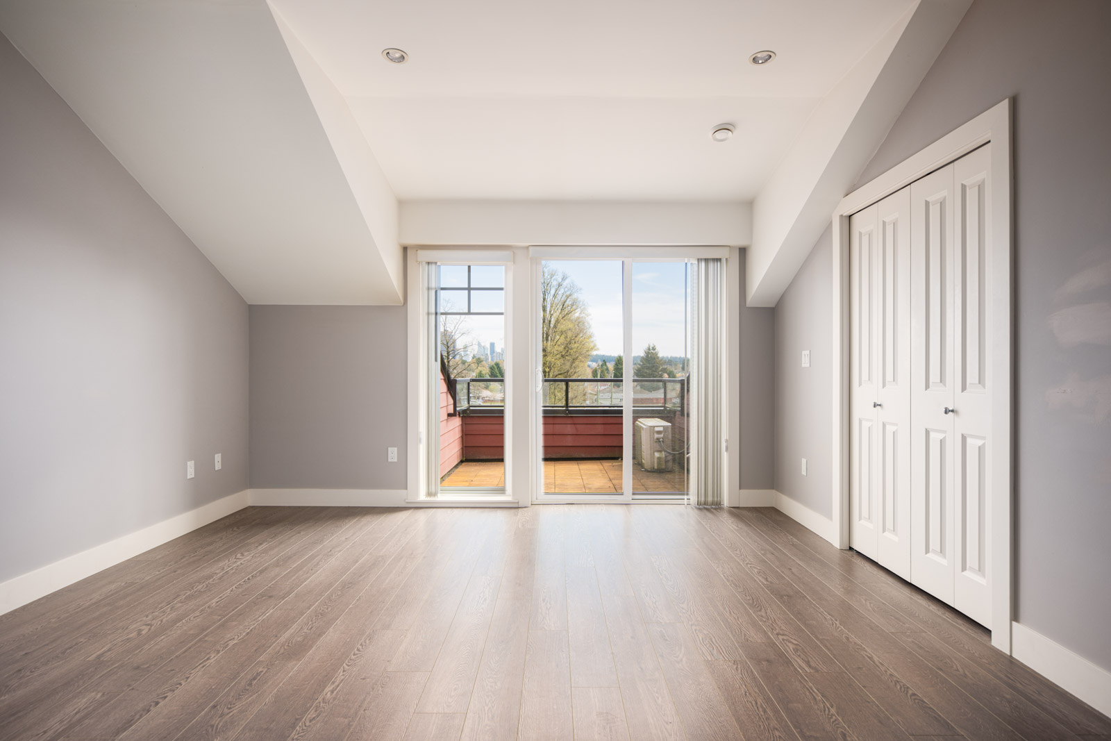 Empty room with gray walls and wood flooring, featuring a closet with double doors and sliding glass doors opening to a balcony with outdoor view.