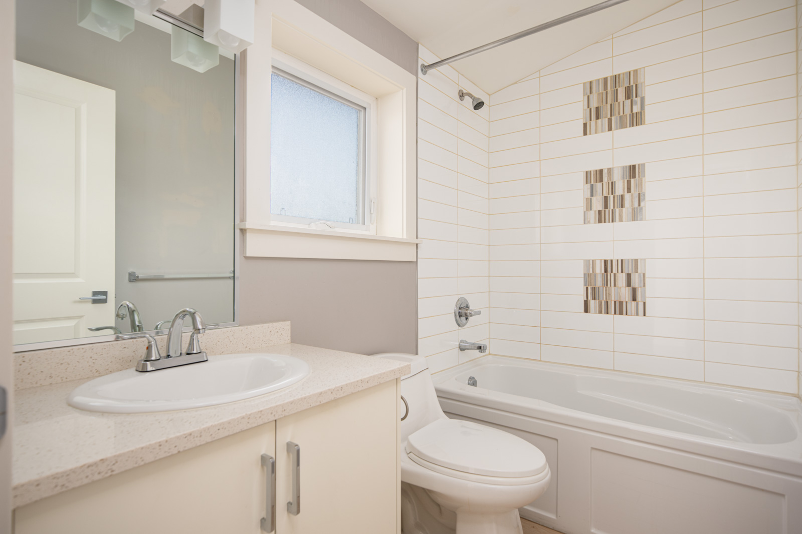 Modern bathroom with a white sink, toilet, bathtub, and tiled shower wall featuring vertical accent tiles, next to a frosted window and mirror.