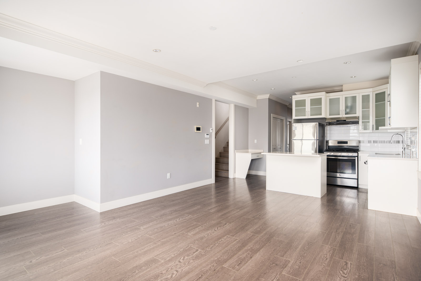 Modern empty kitchen and living area with light gray walls, wooden flooring, stainless steel appliances, and a staircase leading upstairs.