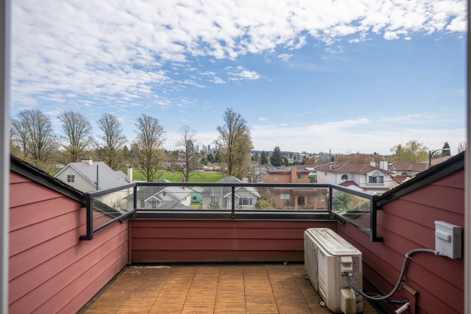 View from a red balcony with glass railing, overlooking rooftops, trees, and a park under a partly cloudy sky. An air conditioning unit is on the balcony floor.