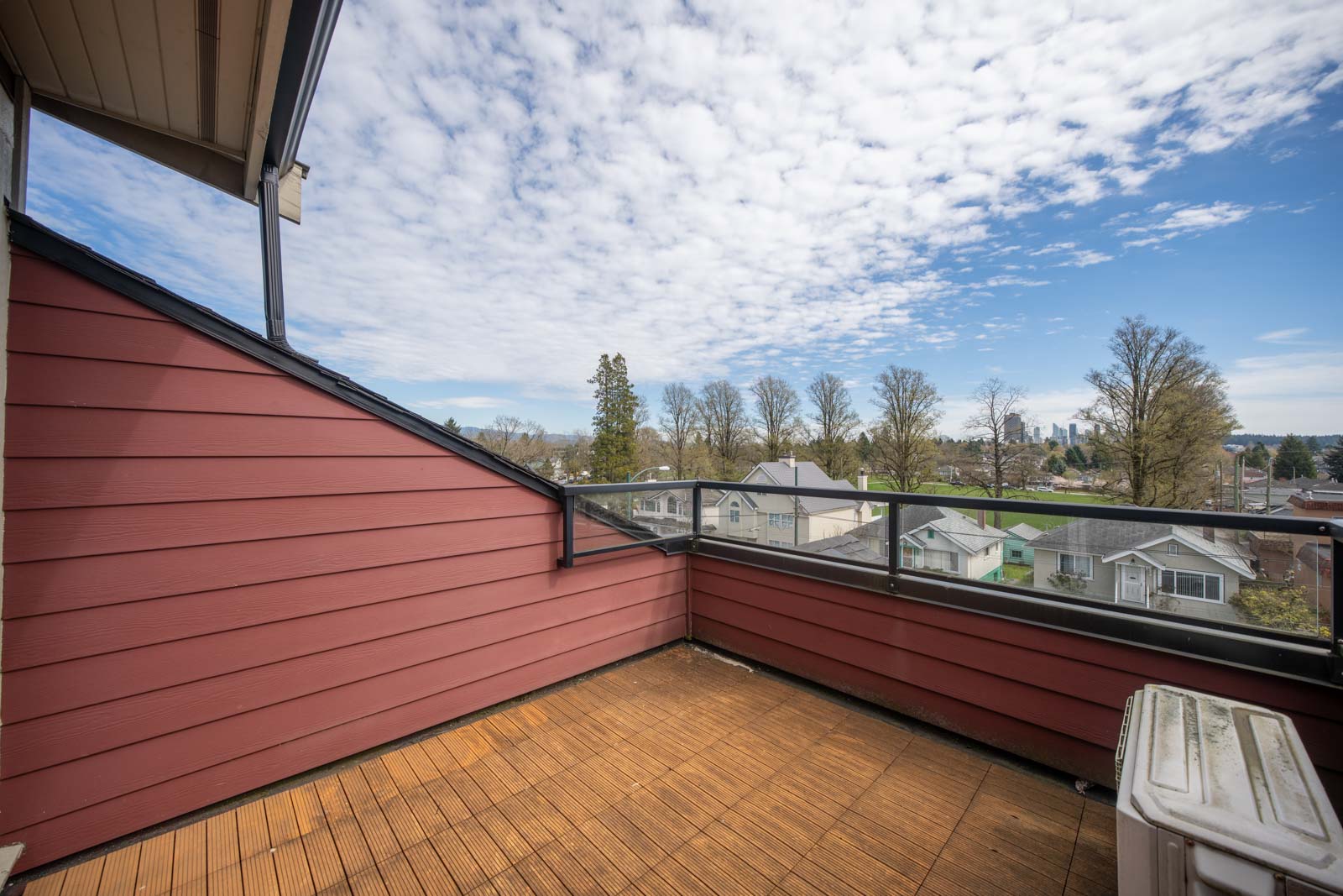 A rooftop balcony with wood flooring and a glass railing overlooks a residential neighborhood under a partly cloudy sky.