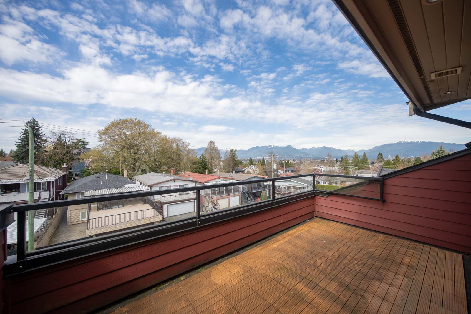 A wooden balcony with glass railing overlooks residential rooftops, trees, and distant mountains under a partly cloudy sky.