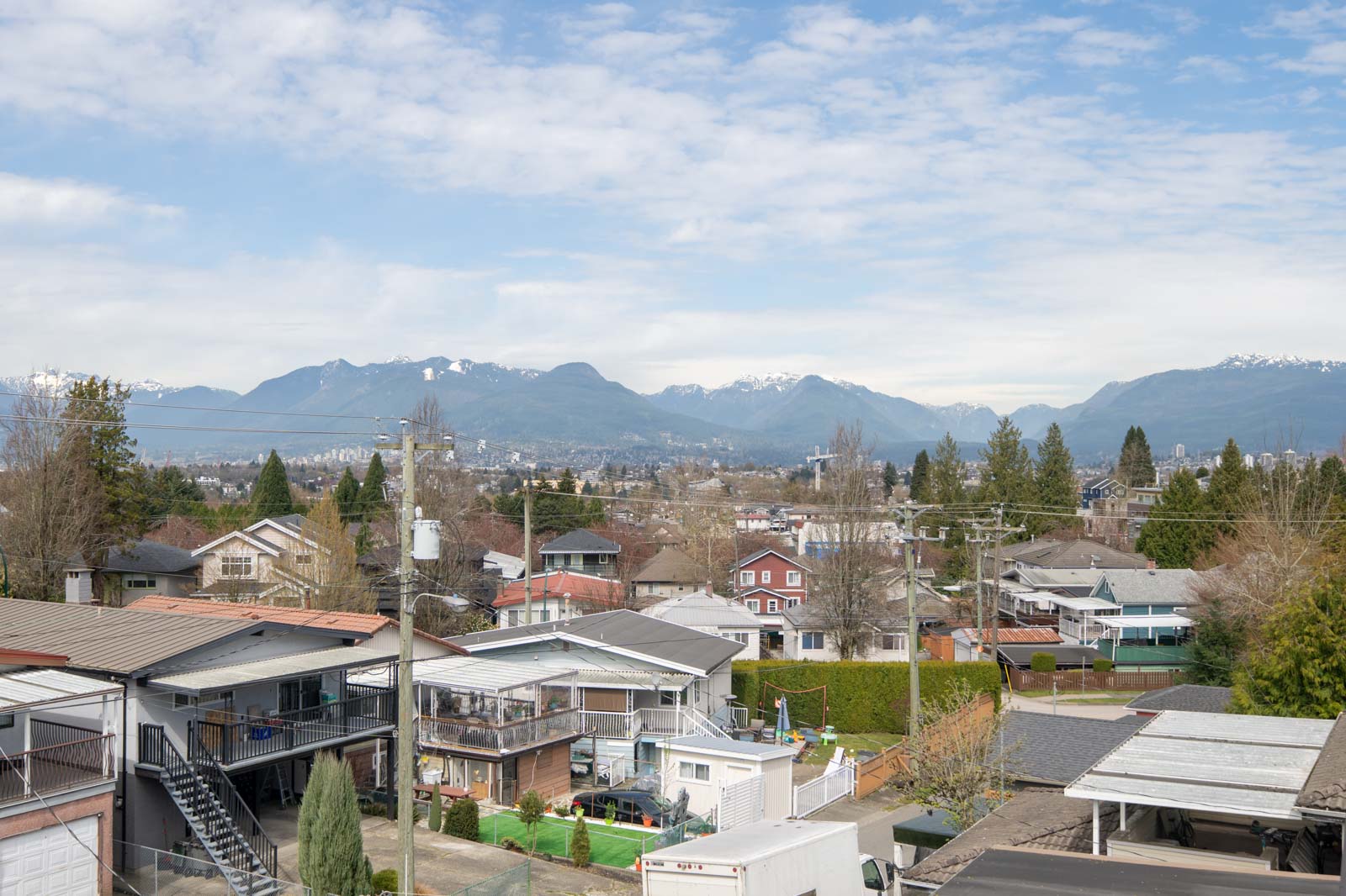 Residential neighborhood with houses, garages, and trees in the foreground, with mountains and a partly cloudy sky in the background.