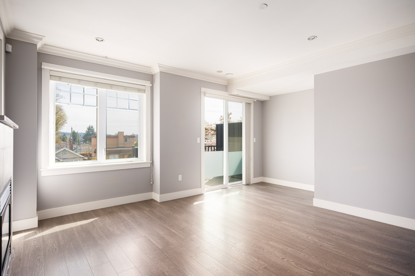 Empty modern living room with large window, sliding glass door to balcony, light gray walls, and wood flooring. Natural light fills the space.