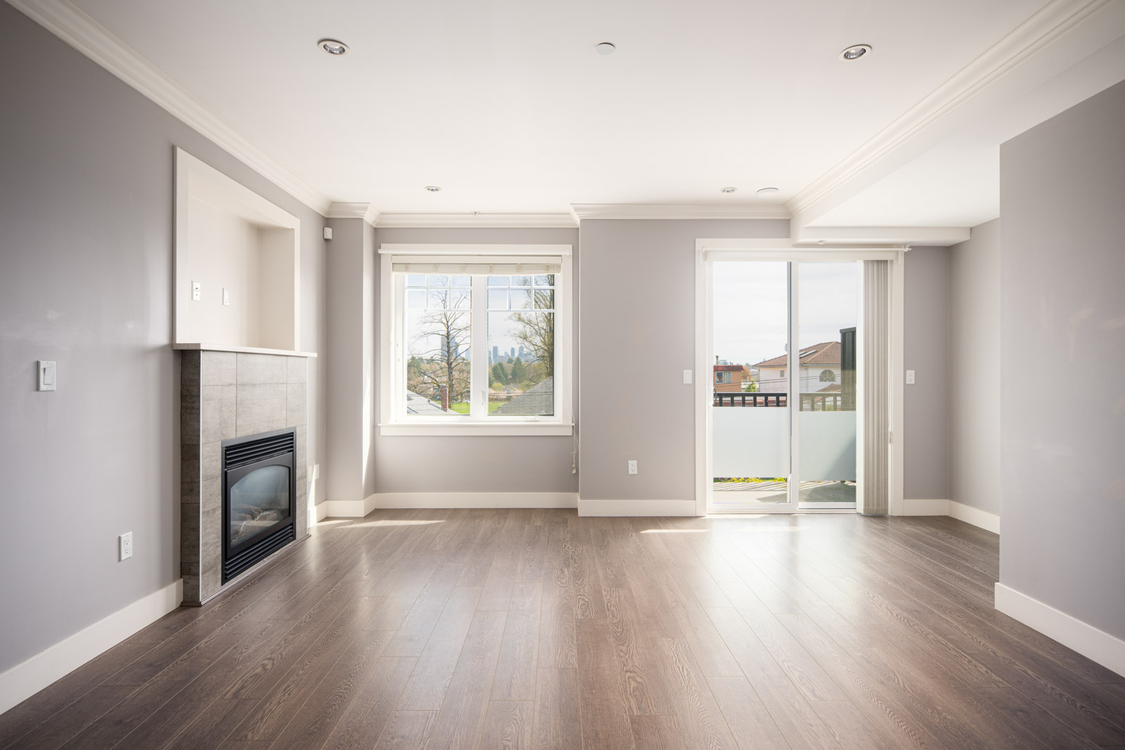 Empty room with light gray walls, wood flooring, a fireplace on the left, a window in the center, and sliding glass doors opening to a balcony on the right.