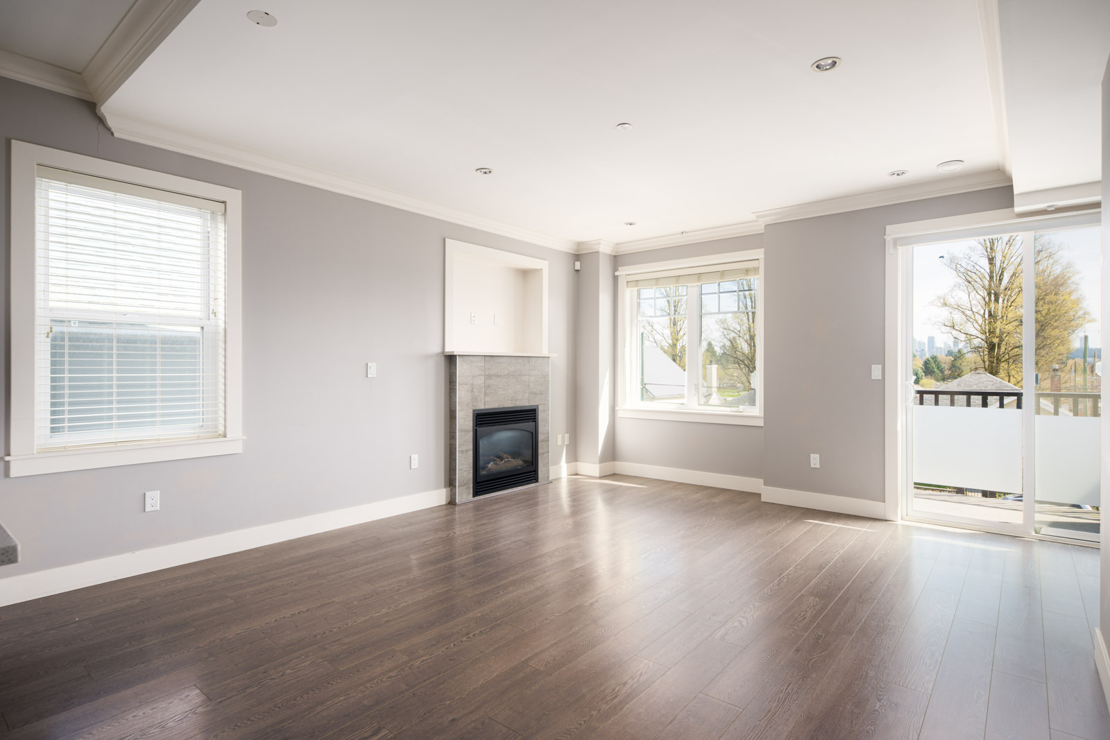 Empty living room with light gray walls, dark wood flooring, a fireplace, two windows, and a glass door opening to a balcony with an outdoor view.
