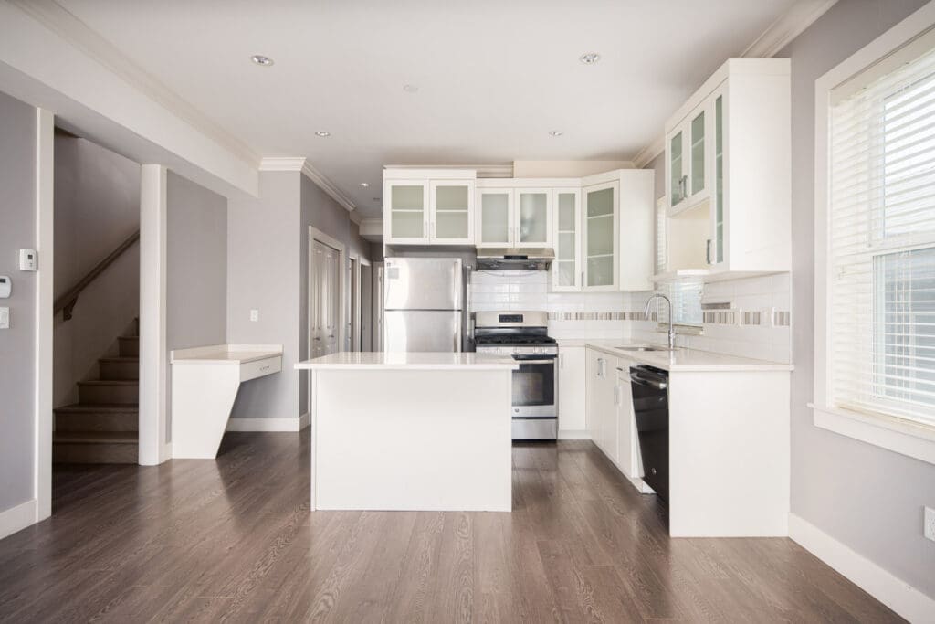 Modern kitchen with white cabinets, stainless steel appliances, an island, dark wood flooring, and a window on the right. Stairs are visible on the left side.