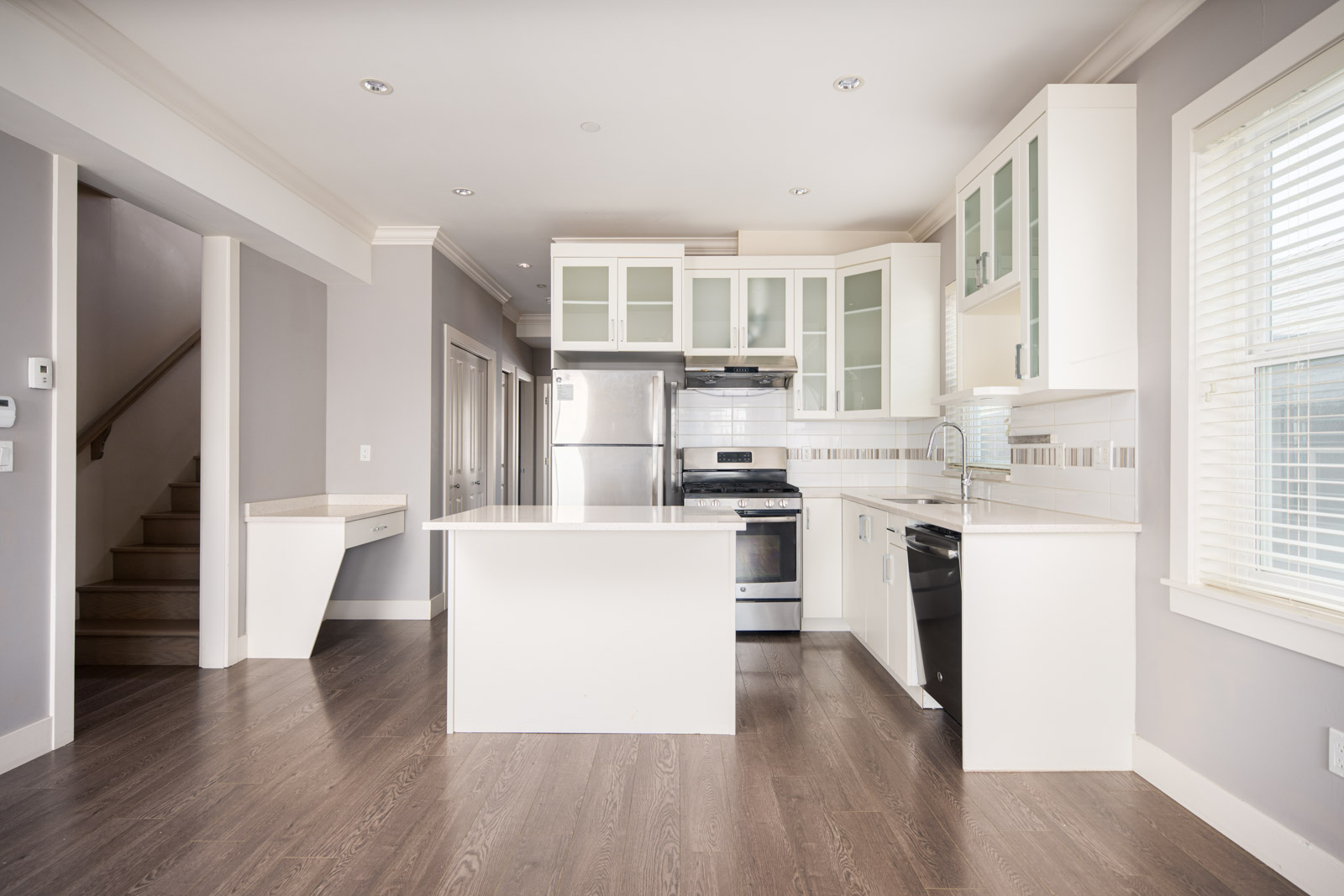 Modern kitchen with white cabinets, stainless steel appliances, an island, dark wood flooring, and a window on the right. Stairs are visible on the left side.