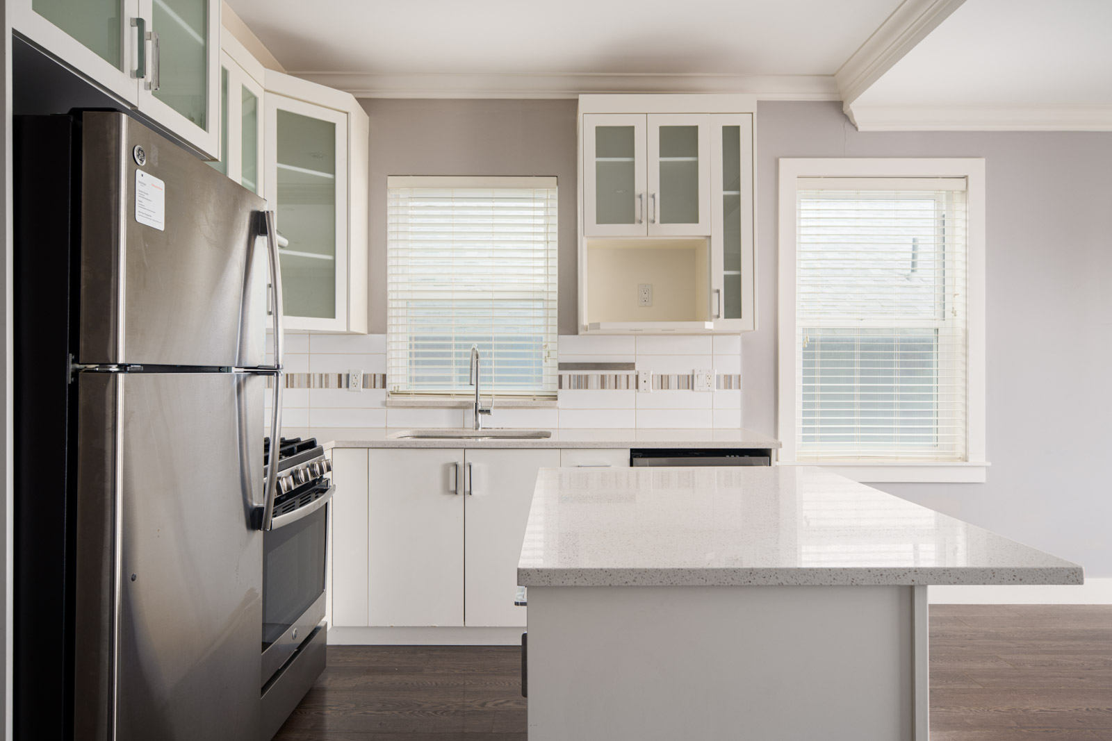 Modern kitchen with stainless steel refrigerator, white cabinets, gas stove, large island, and windows with blinds allowing natural light.