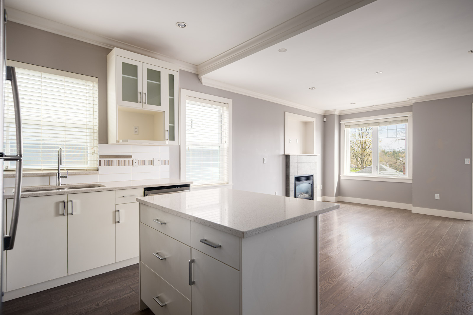 Modern, unfurnished kitchen with white cabinets, island counter, stainless steel appliances, and large windows leading into an open living space with wood flooring.