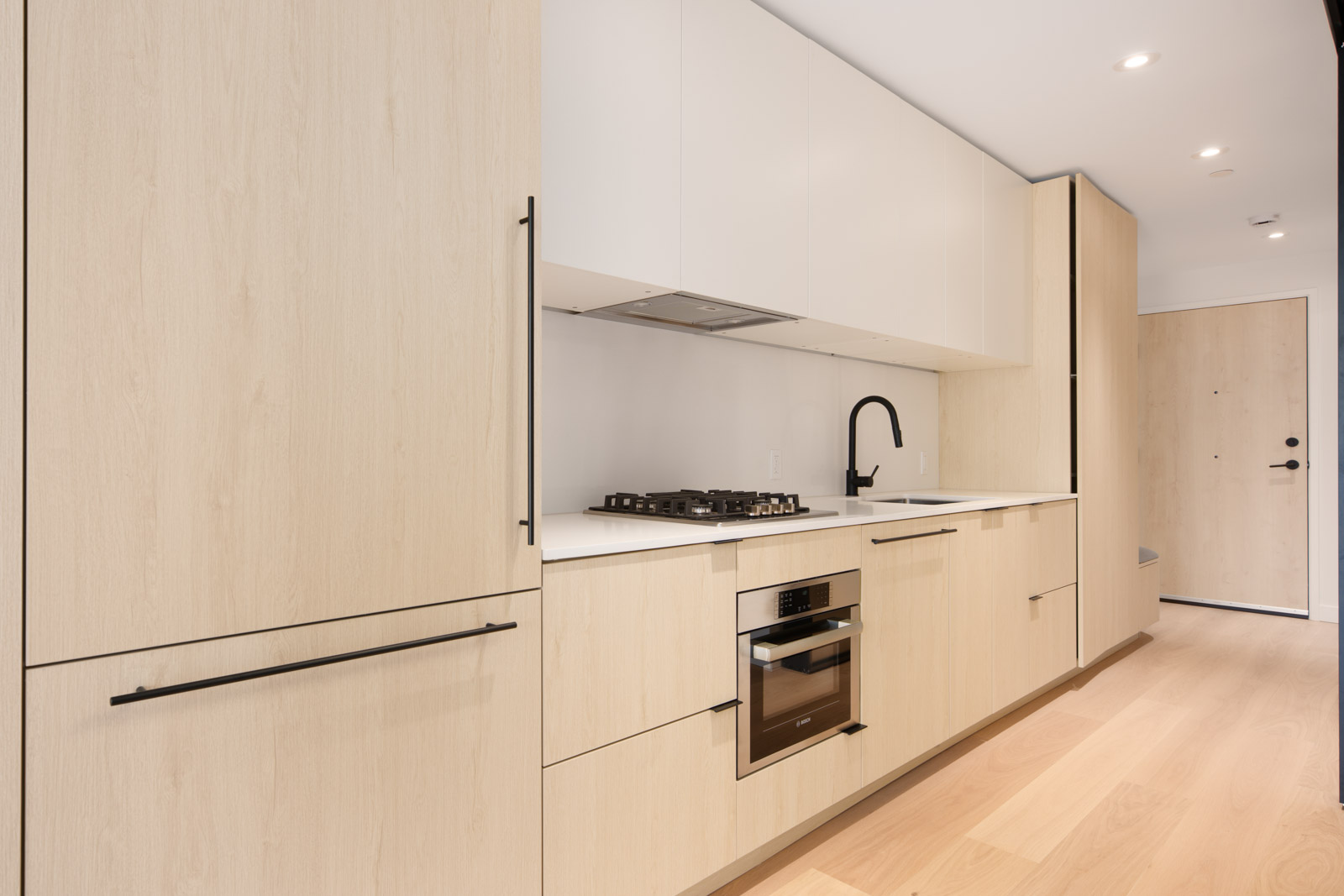Modern kitchen with light wood cabinets, white countertops, a built-in oven, gas cooktop, black faucet, and minimalist design.