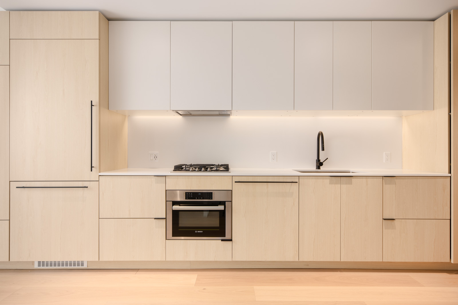 Modern kitchen with light wood cabinets, white countertops, a built-in oven, gas stovetop, black faucet, and integrated appliances under bright, even lighting.