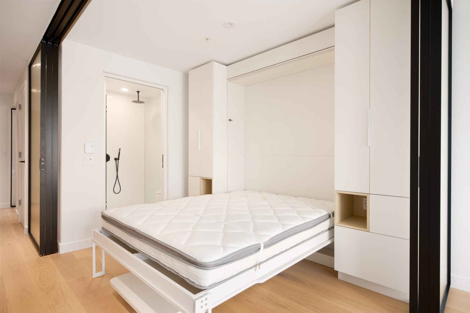 A modern, minimalistic room with a Murphy bed extended, white built-in cabinets, light wood flooring, and an open bathroom with a walk-in shower in the background.