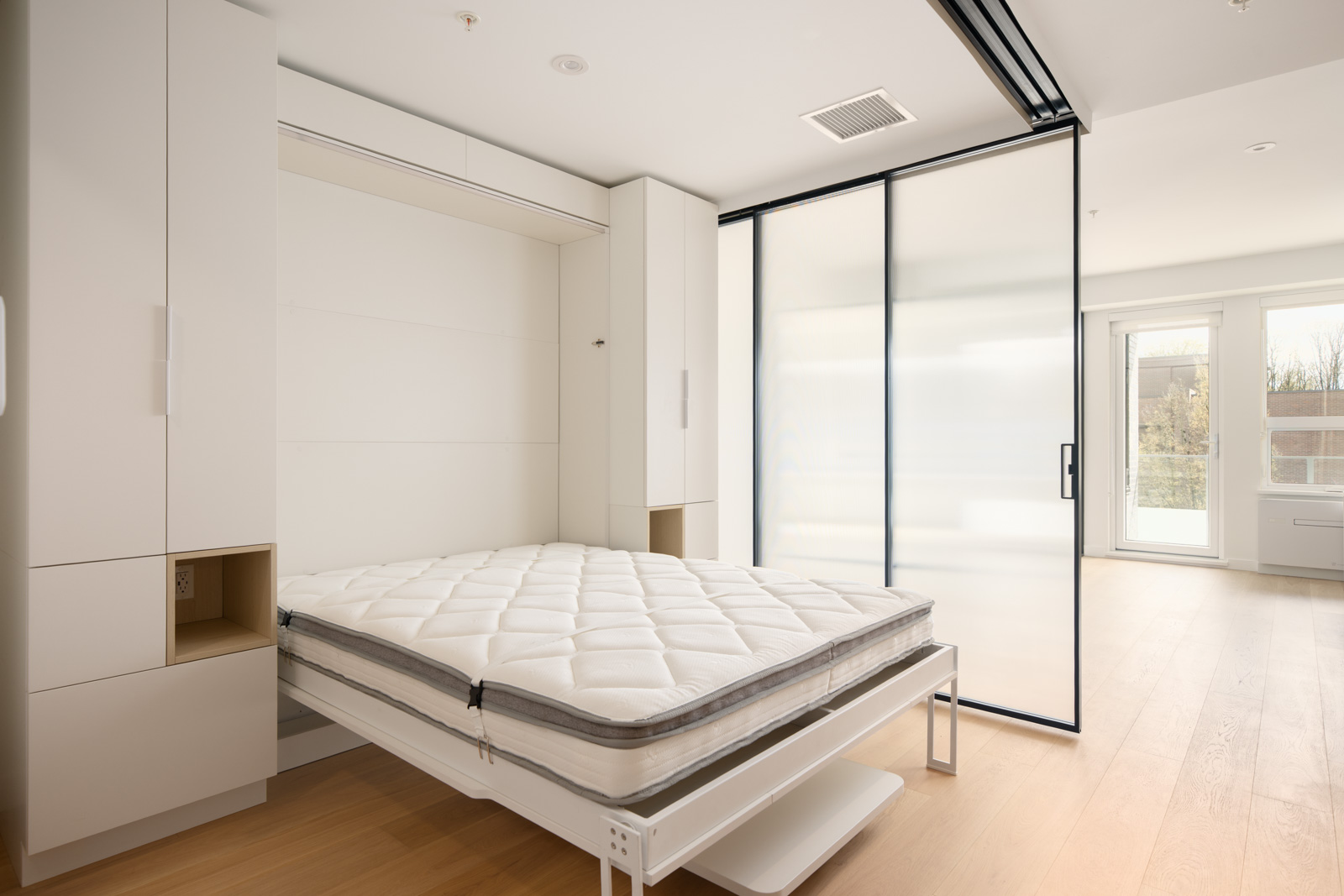 A modern, minimal bedroom with a pull-down Murphy bed, built-in white storage cabinets, and frosted sliding glass doors leading to a bright room with large windows.