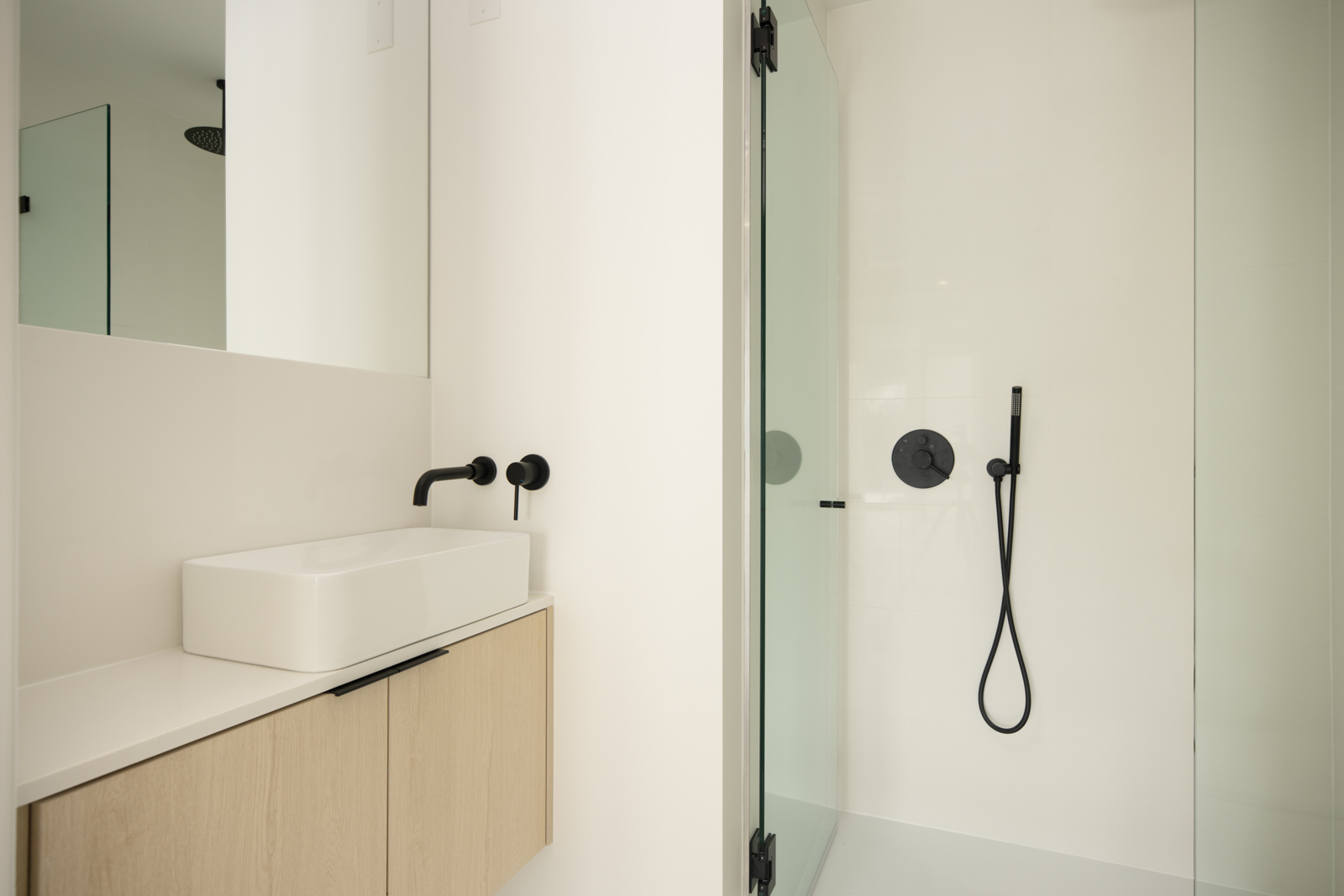 Minimalist bathroom with a floating wooden vanity, rectangular sink, black faucet, large mirror, and a glass shower with black fixtures.