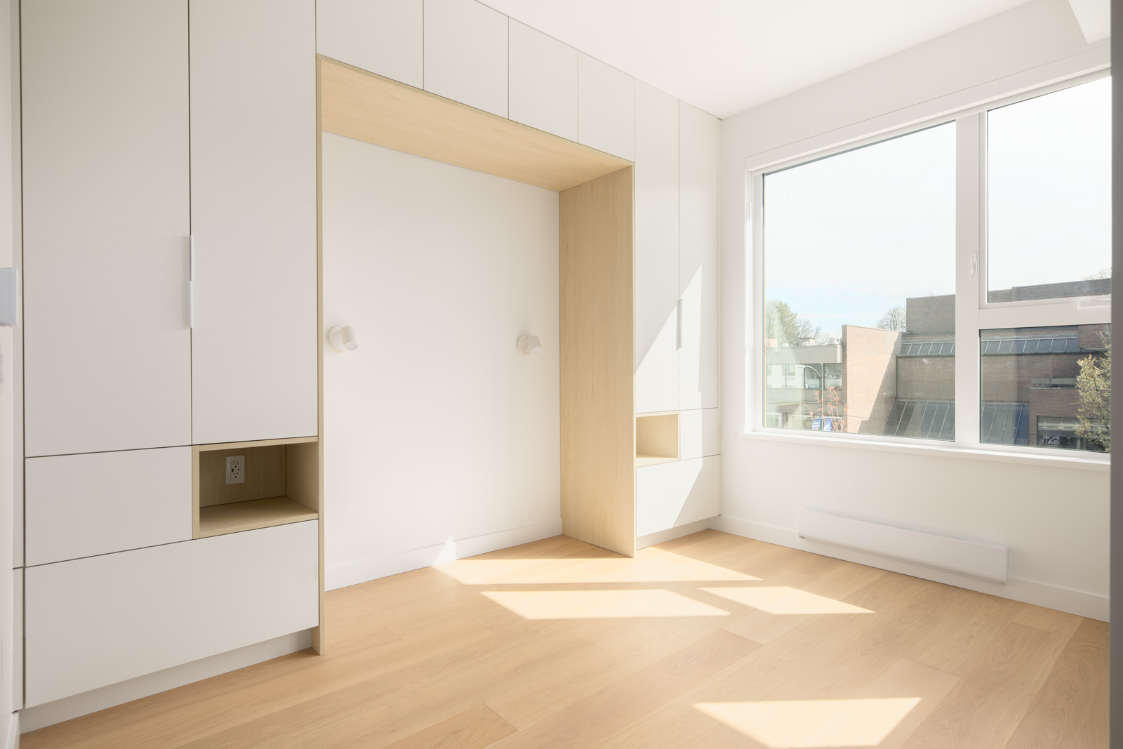 Minimalist, unfurnished bedroom with built-in white cabinets, light wood accents, and a large window letting in natural light, overlooking a building outside.
