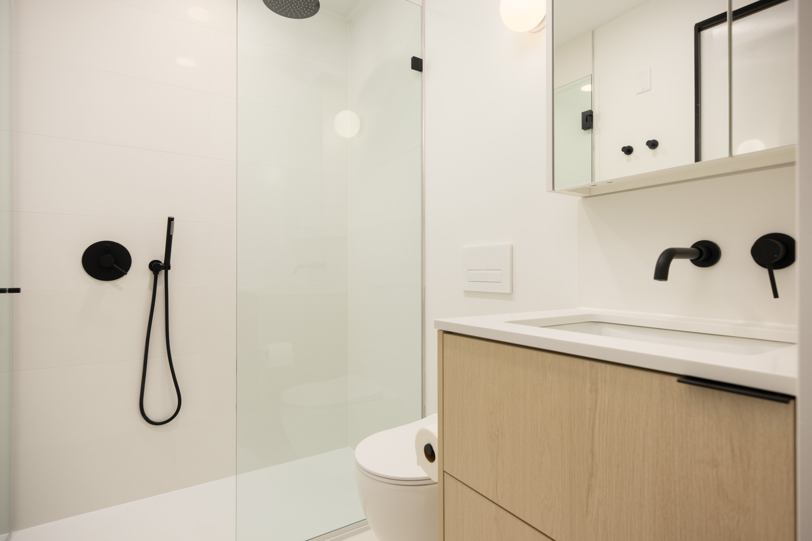 Modern bathroom with glass shower, black fixtures, light wood vanity, white countertop, wall mirror, and minimal decor.